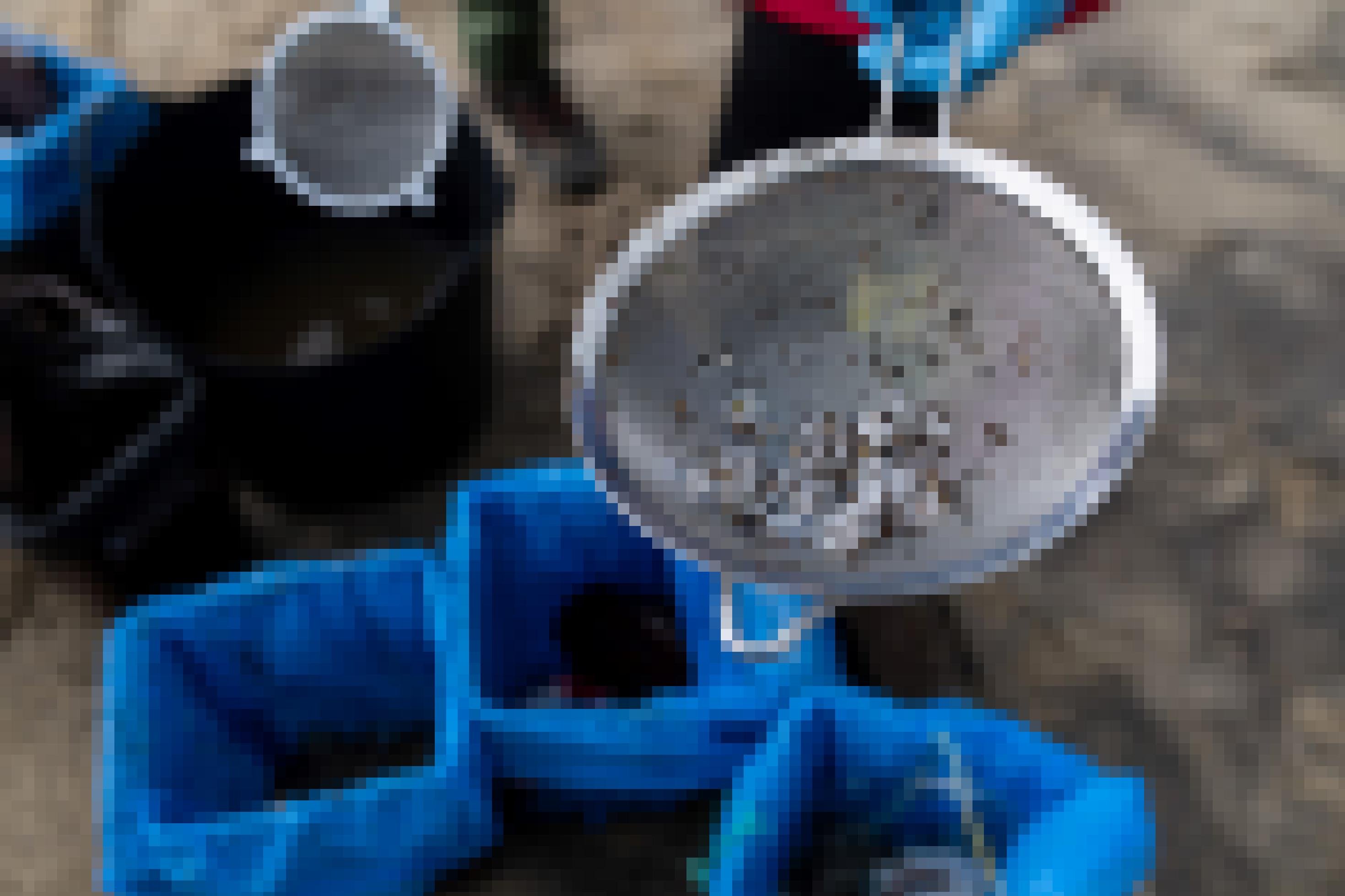 Zwei Küchsensiebe werden in die Kamera gezeigt. An den Sieben hängen kleine weiße Plastikkügelchen, vermischt mit etwas Sand. Der Hintergrund zeigt den Sandboden, auf dem die Helfer stehen, sowie drei Mülleimer.