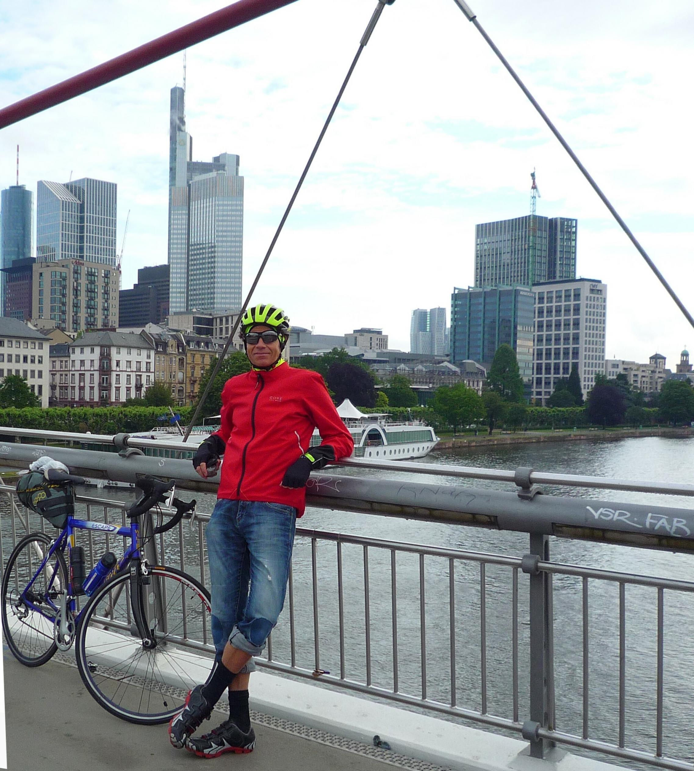 Martin C Roos steht mit seinem Rennrad am Geländer des Holbeinstegs, hinter ihm der Main vor der Skyline Frankfurts.