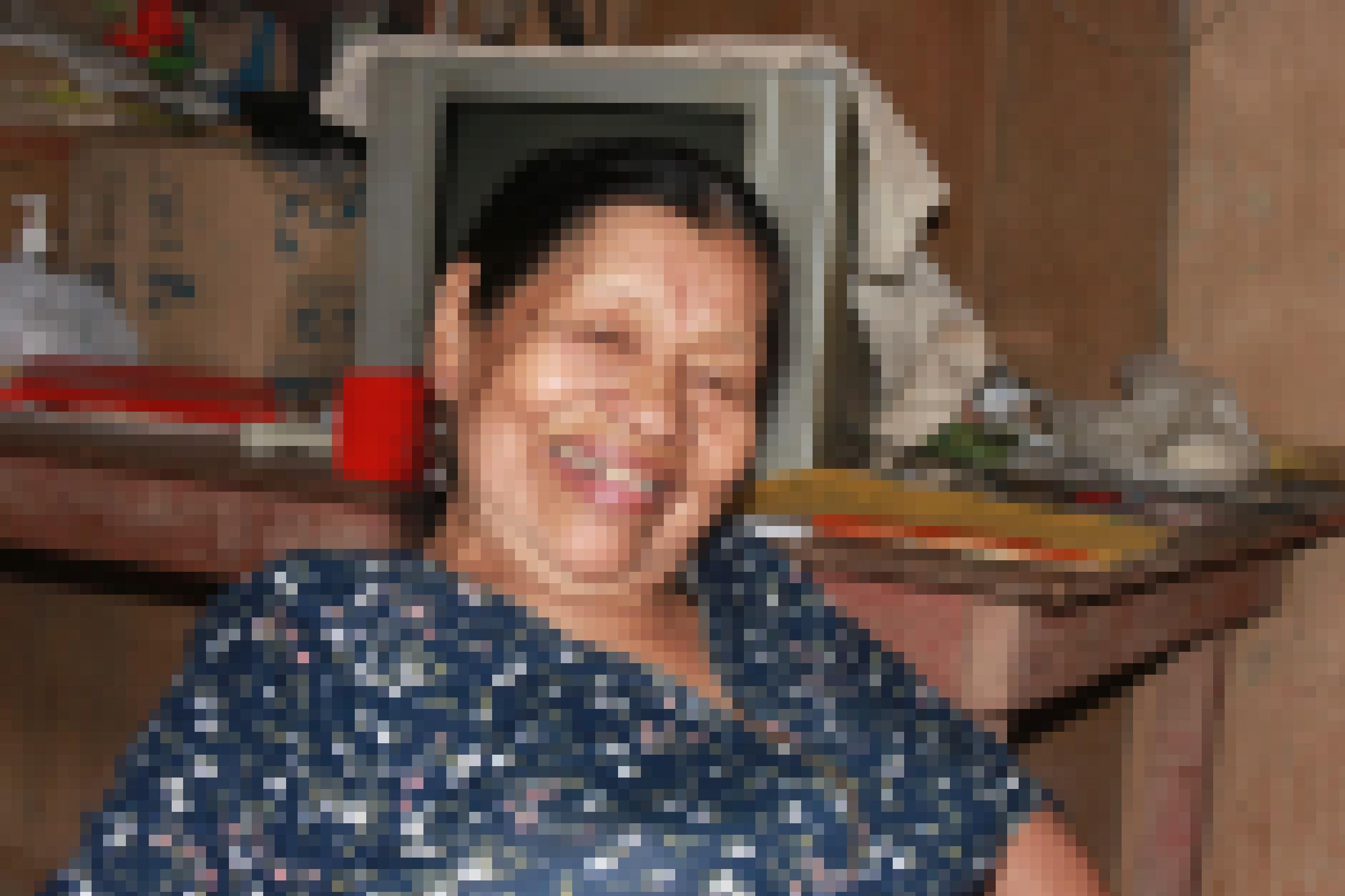 Brustbild von Frau mit runzligem Gesicht, schwarzen zurückgekämmten Haaren, lacht  in die Kamera, im Hintergurnd ein altes PC_Modell