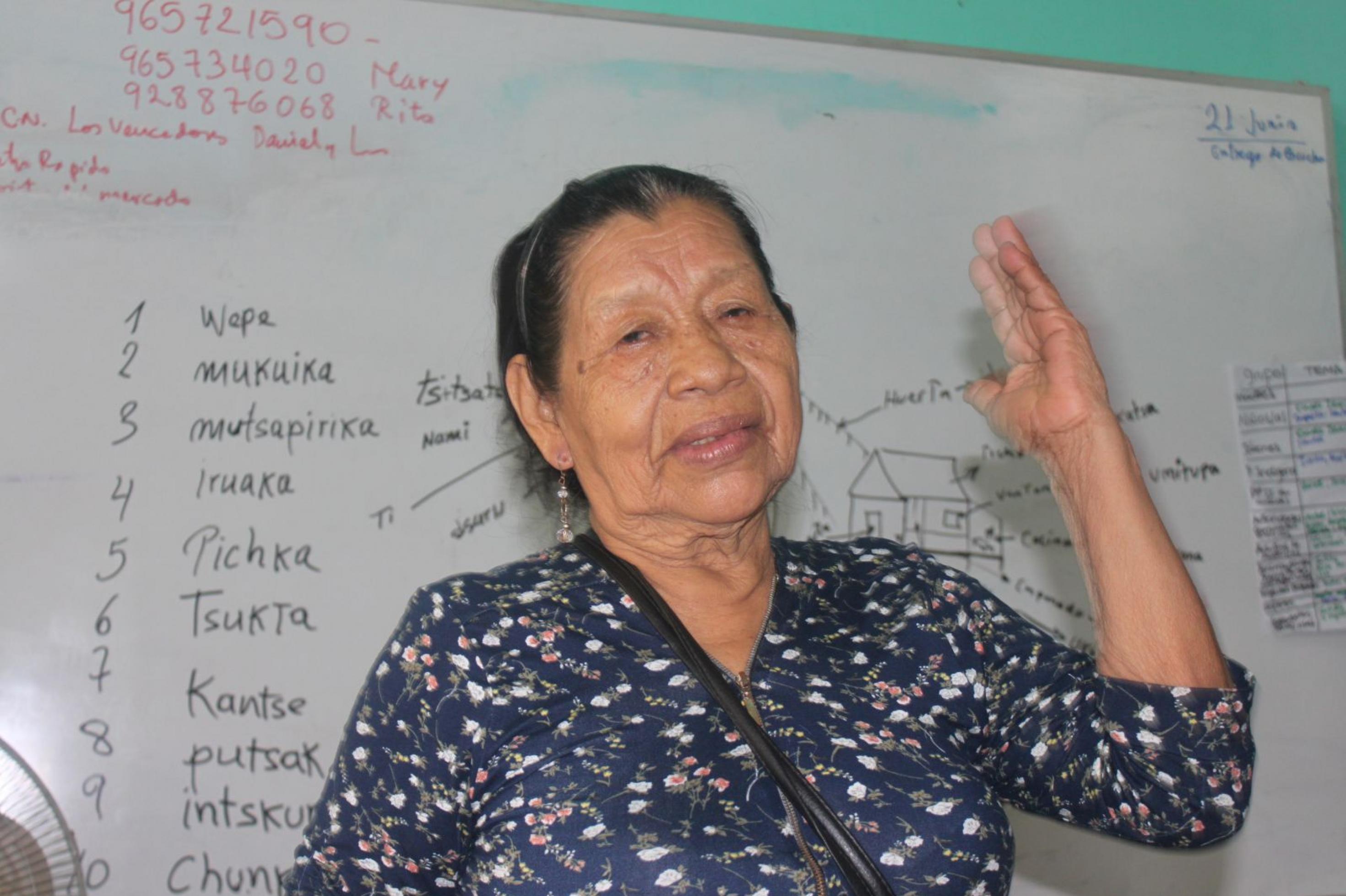 Alte Frau mit runzligem Gesicht, schwarzem, zurückgekämmtes Haar, steht vor einem Whiteboard, auf dem Wörter in der Kukama-Sprache stehen. Sie gestikuliert mit ihrer Hand.