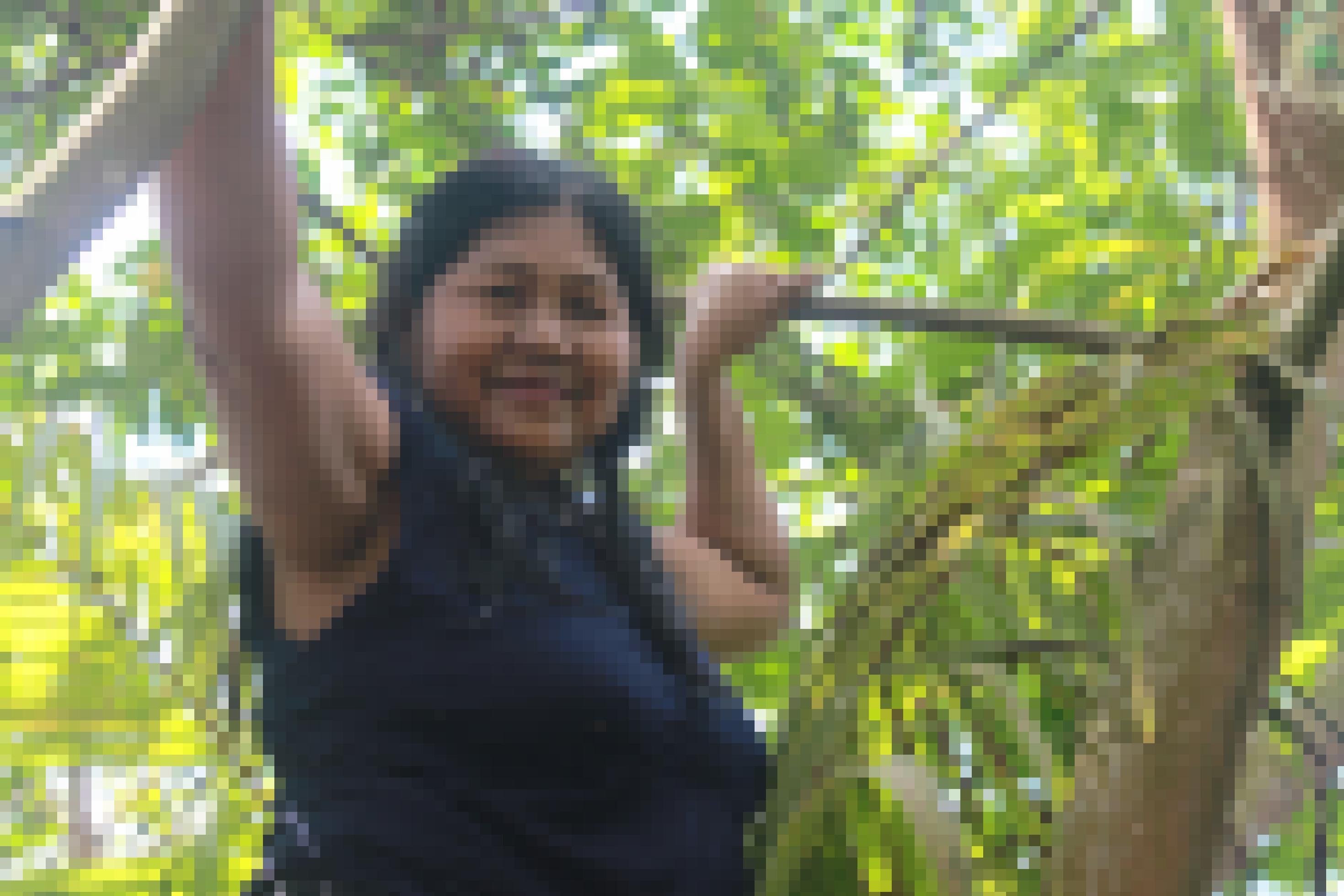 Frau mit brauner Haut, mittelalt, schwarze Haare bis auf die Schultern, ärmelloses blaues T-Shirt, blickt von einem Baum herunter in die Kamera.