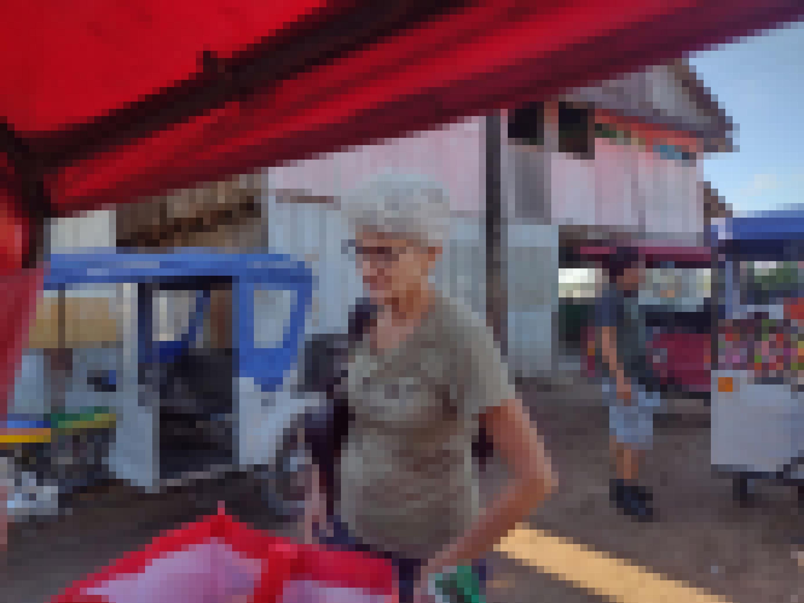 Mehrere Tuktuks. Frau mit weissen kurzen Haaren und Brille steht hinter einem Tuktuk und schaut auf eine Tasche vor ihr.