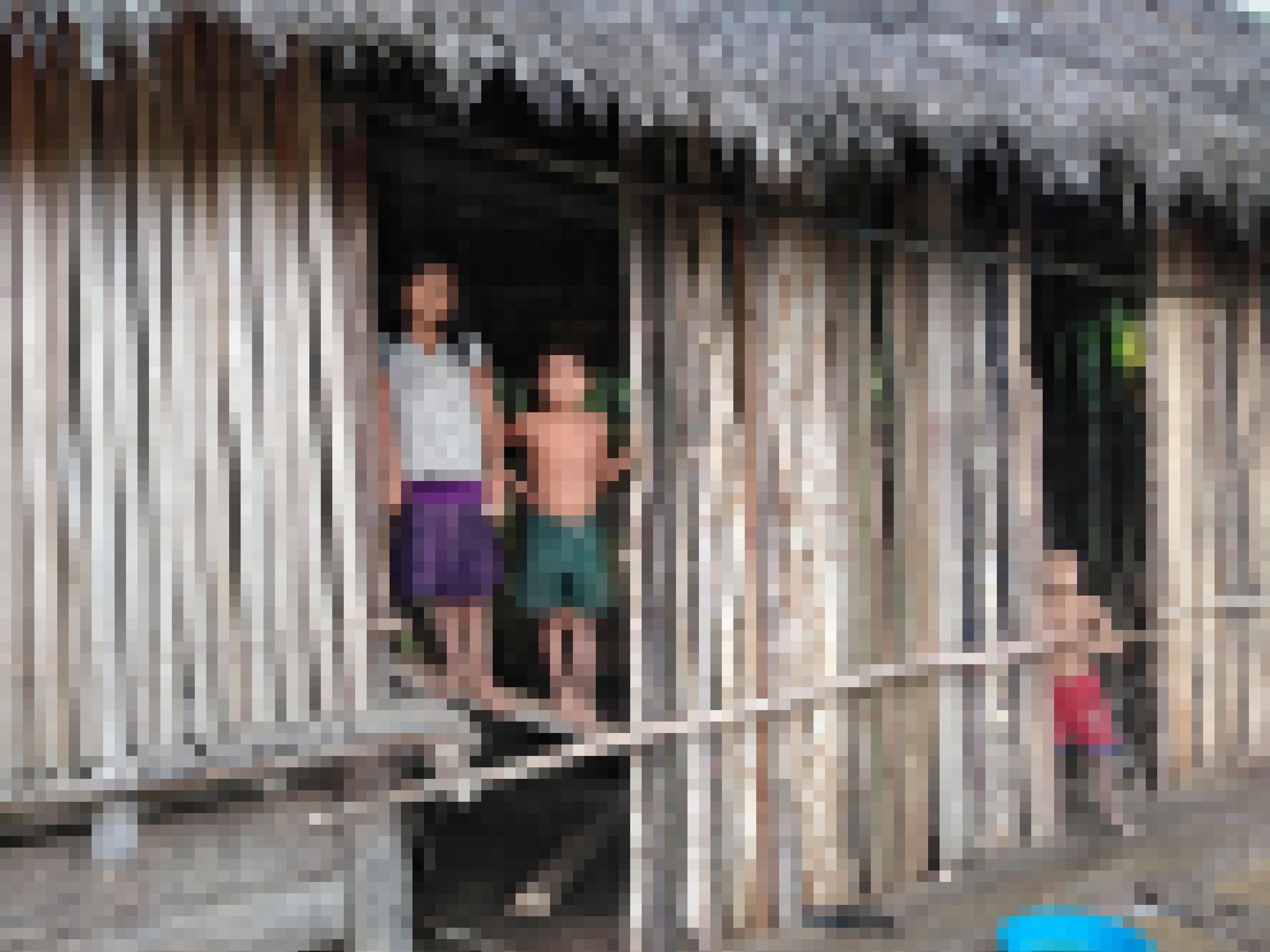 Ein Haus aus Pfählen und Palmdach, daraus schauen ein ca. 12-jähriges Mädchen und ein ca. 6-jähriger Junge, beide mit indigenen Gesichtszügen.