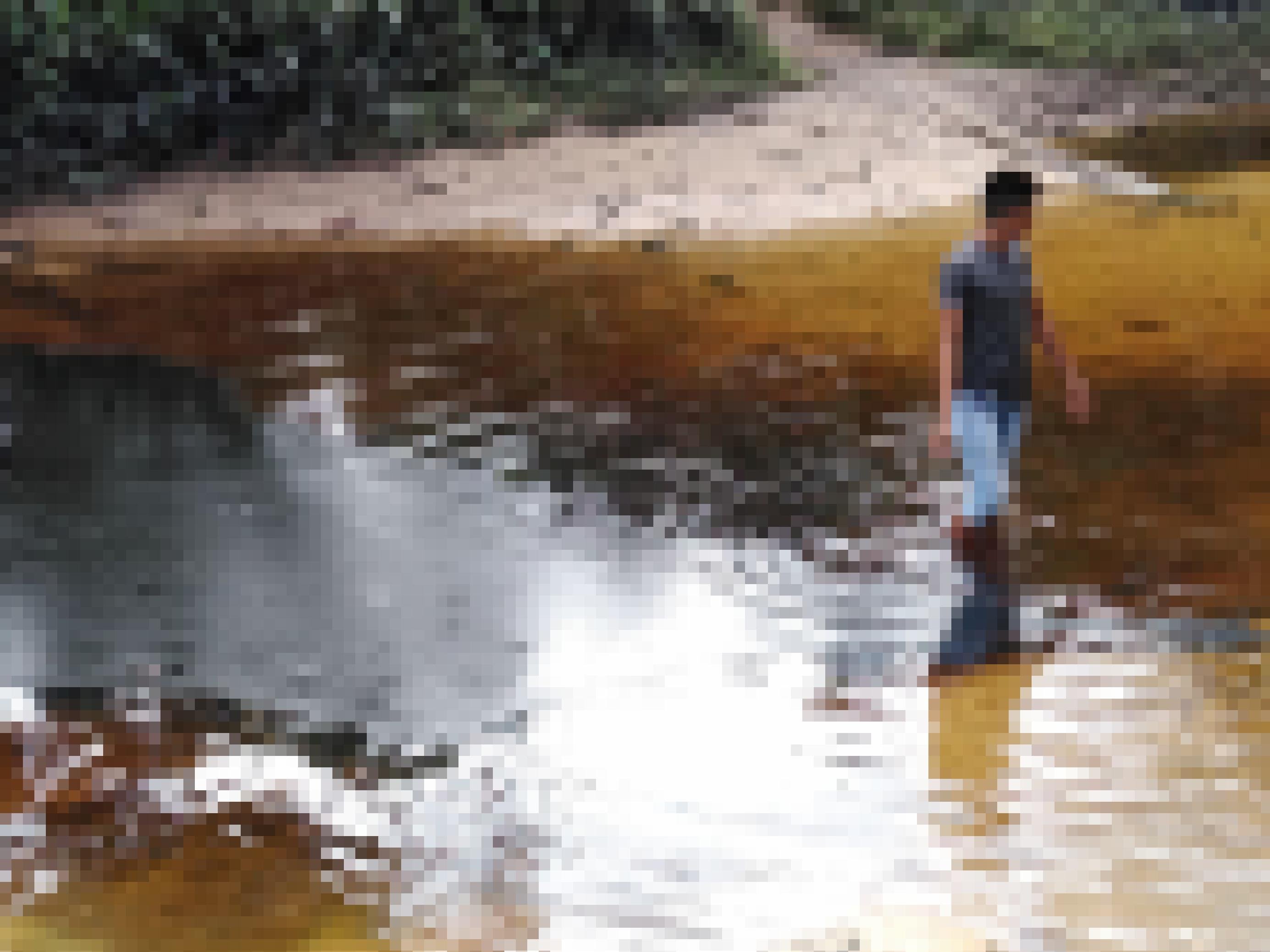 Ufer eines kleinen Flusses, am Ufer eine grüne Böschung. Das Wasser im Fluss ist dunkel- bis hellbraun vom Erdöl. Auf der rechten Seite des Bildes watet ein junger Mann mit hochgekrempelten Hosen durch das Wasser.
