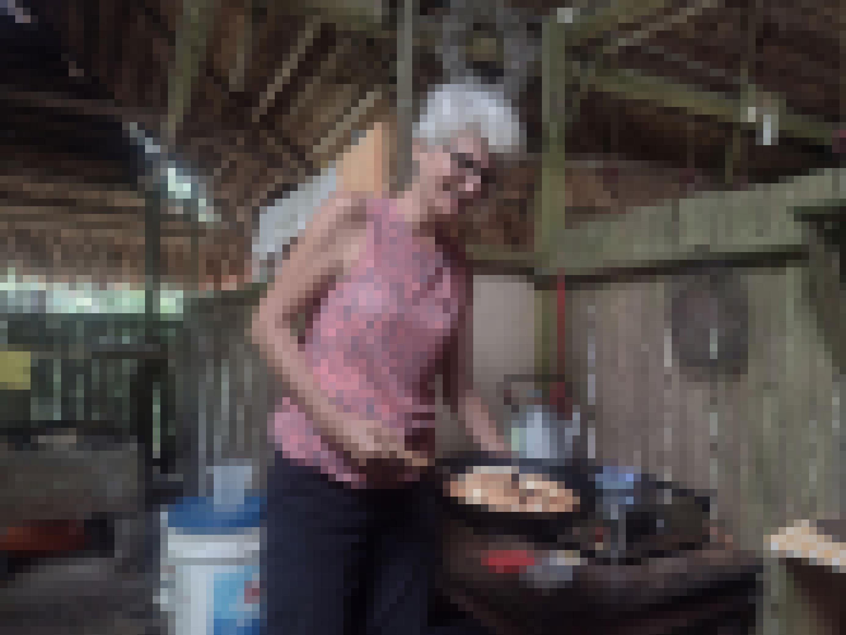 Eine weisse Frau mit weissen kurzen Haaren und Brille toastet Brot in einer Pfanne an einem Gaskocher. Sie befindet sich in einem Haus mit offenen Holzwänden und einem Palmendach.