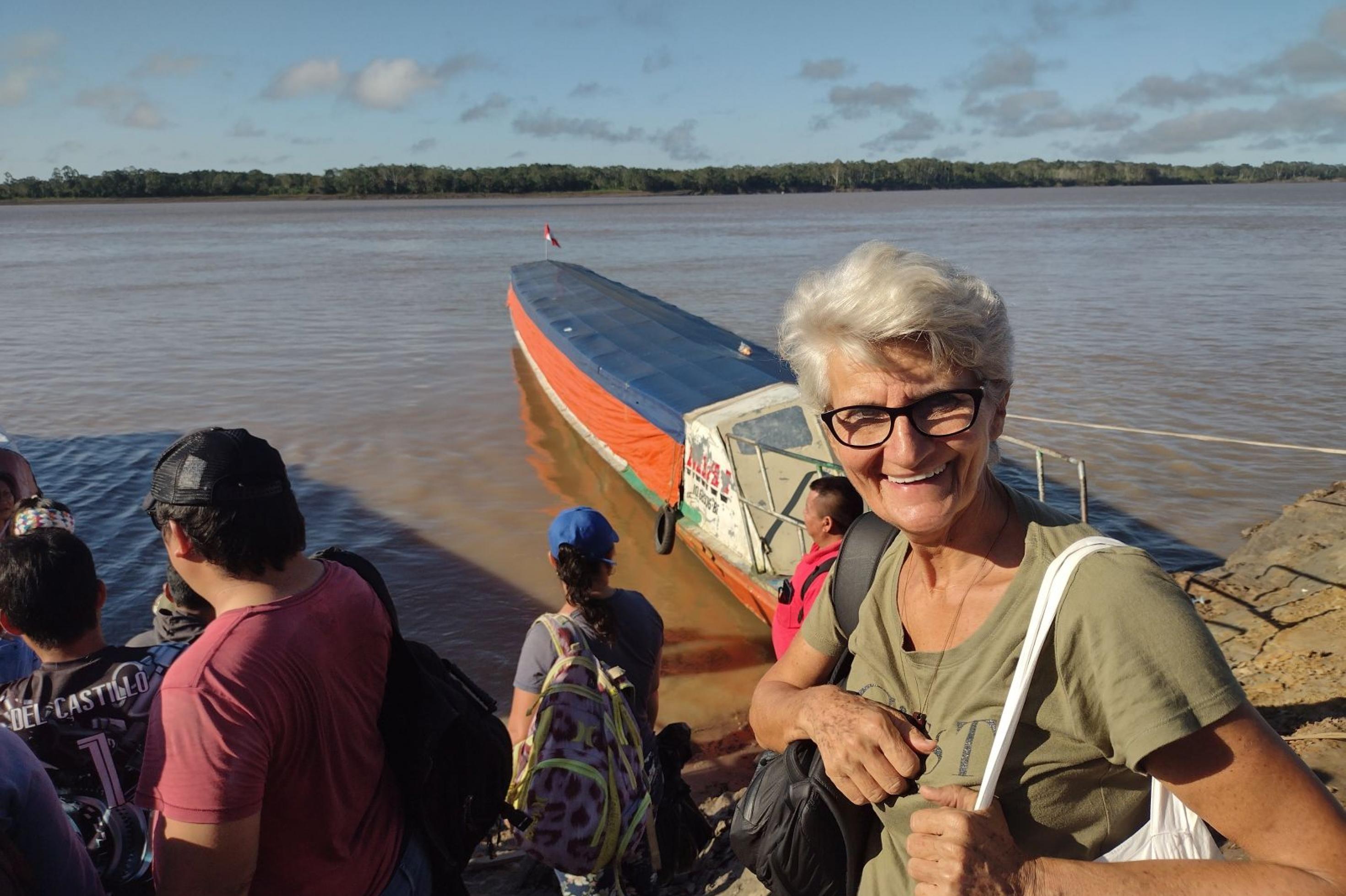Am Fluss, mehrere Boote legen an, davor lacht eine Frau mit kurzen weissen Haaren, Brille und zwei Stofftaschen über den Schultern in die Kamera.