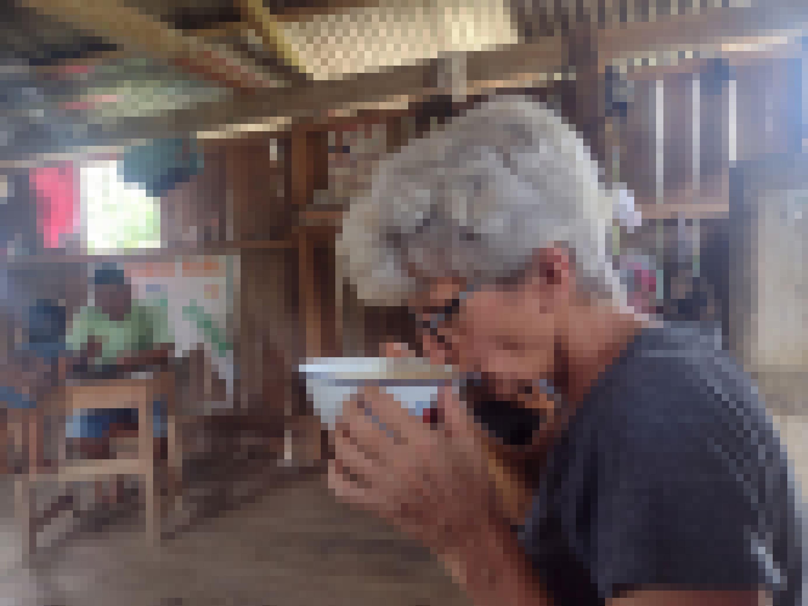 Im Vordergrund eine Frau mit kurzen weissen Haaren und Brille, trinkt ein weisses Gebräu aus einer breiten Schale. Im Hintergrund ein Mann an einem Tisch in einer Holzhütte, davor ein Kind.