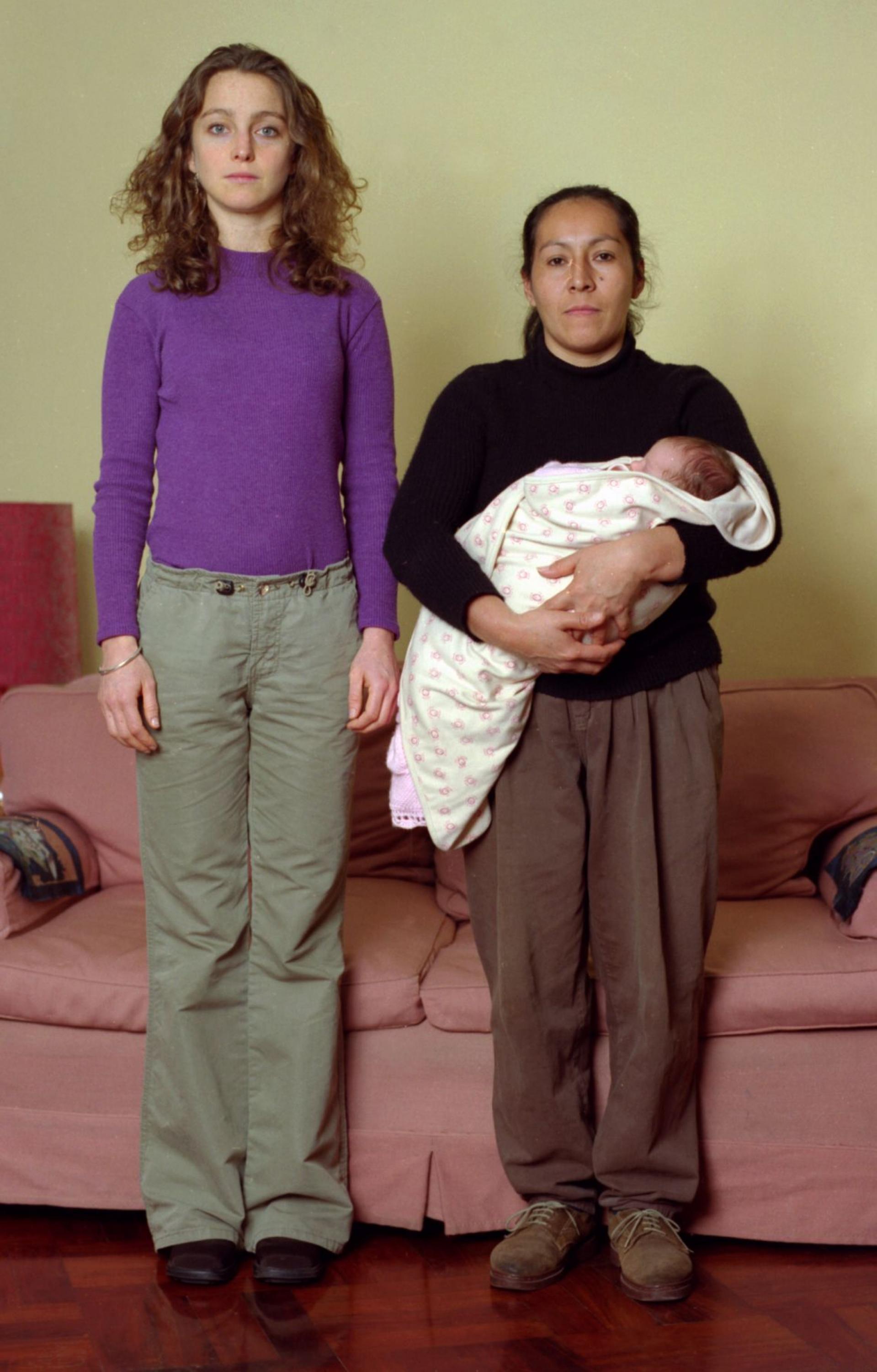 Zwei Frauen stehen nebeneinander und schauen in die Kamera. Links eine junge schmale Frau mit blonden Locken, rechts eine kleinere, aeltere, braunhaeutige Frau mit einem Baby im Arm.