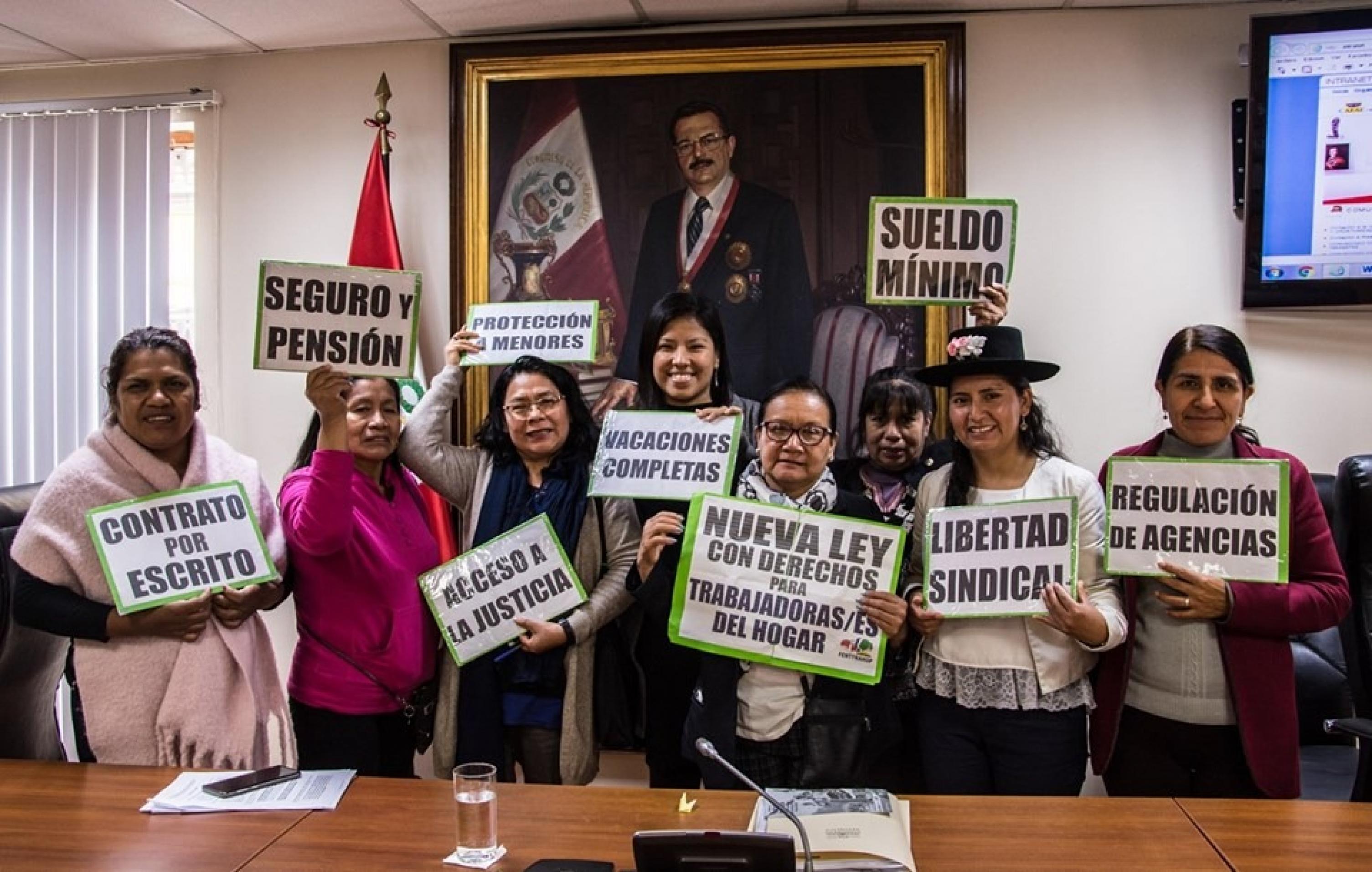 Acht Frauen in einem Raum, heben Plakate mit Forderungen nach besseren Bedingungen fuer Hausangestellte in die Kamera. Im Hintergrund das Oelgemaelde eines peruanischen politischen  Wuerdentraegers.