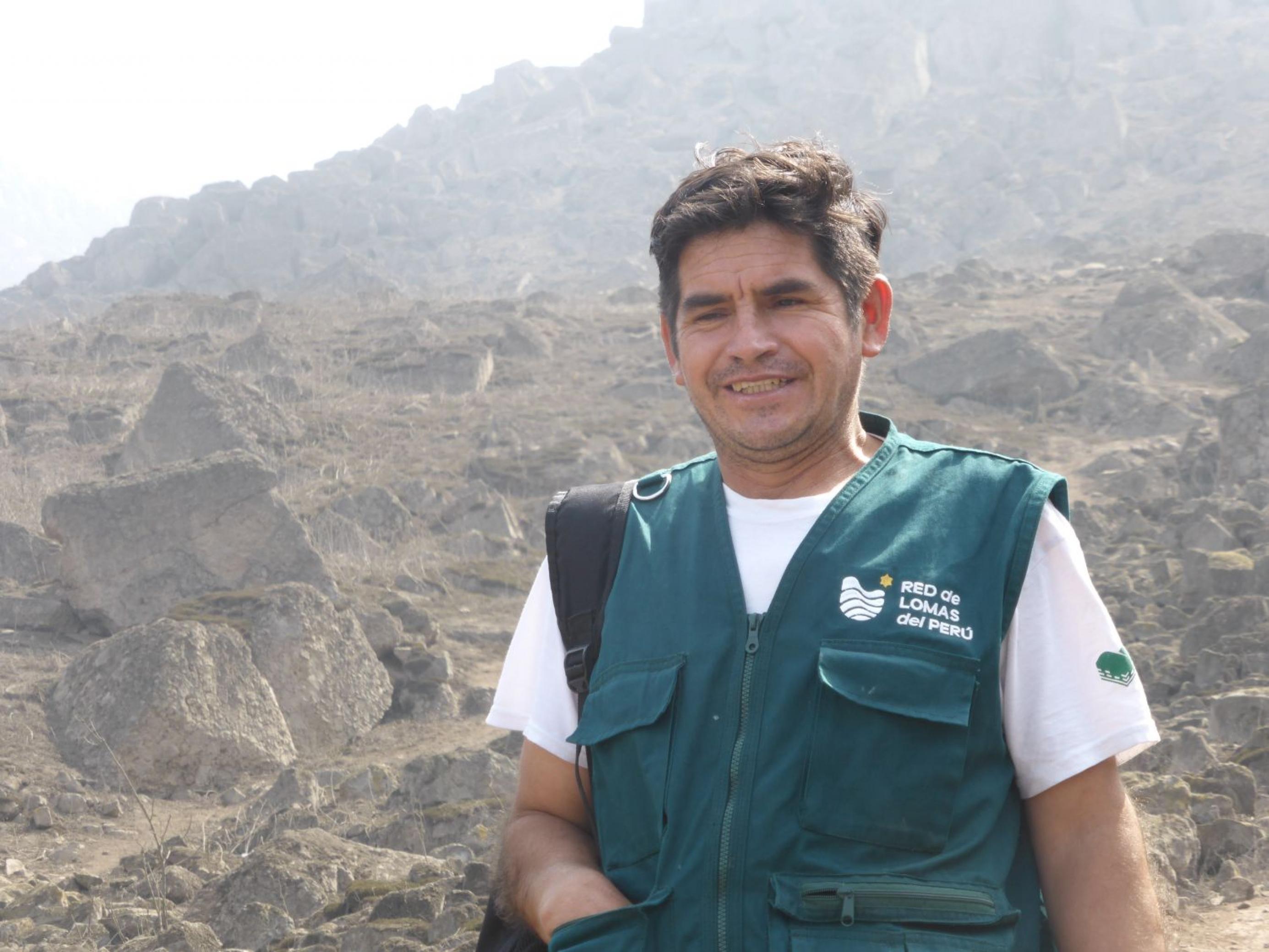 Mann, ca. 40 Jahre, mit kurzärmligem weissen T-Shirt und grüner Jacke, im Hintergrund eine bergige Steinlandschaft
