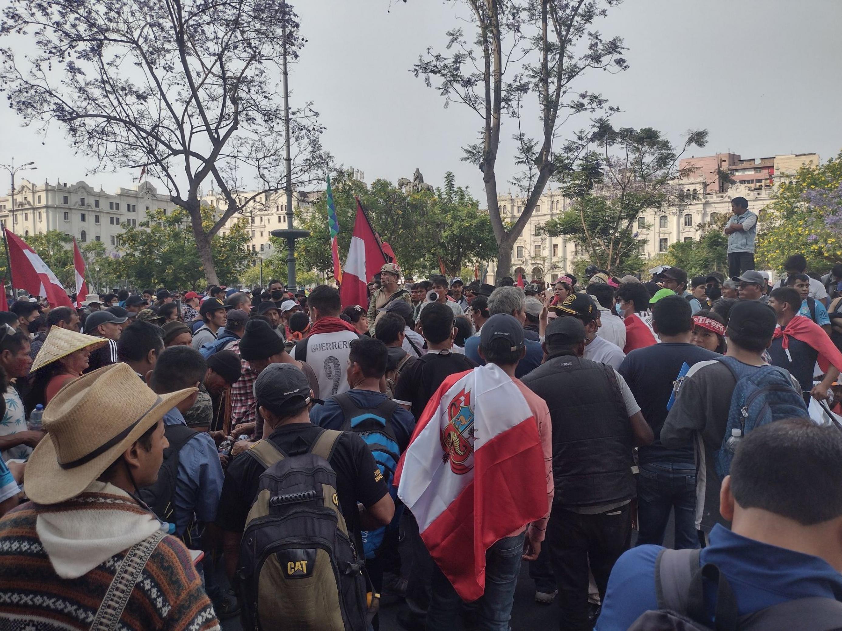 Menschenansammlung vor einem öffentlichen Platz. Einige tragen die peruanische Flagge auf dem Rücken. Sie lauschen einem Mann, der auf einer Bank steht.
