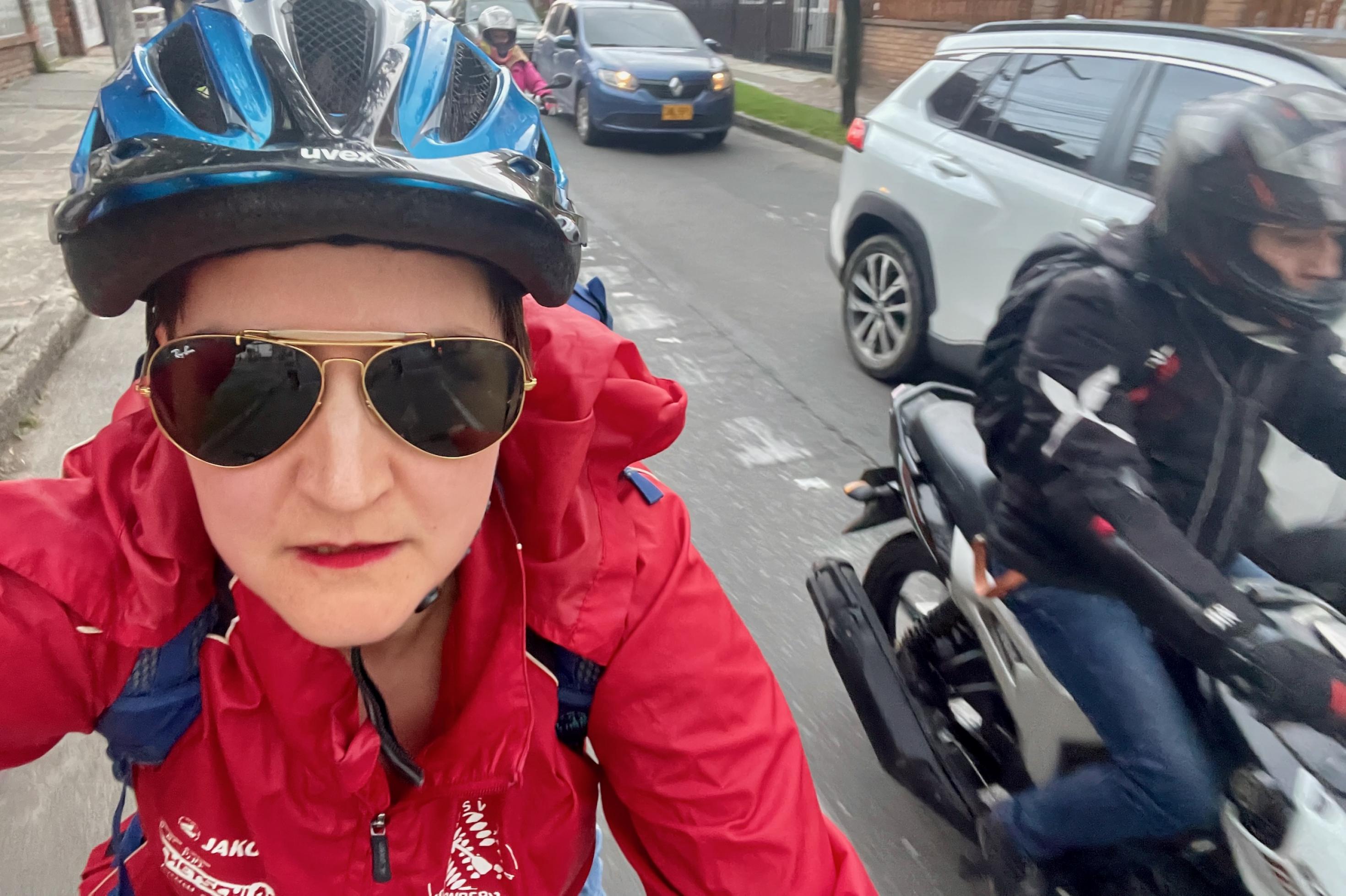 Die Autorin in der roten Radlerjacke und mit blauem Helm blickt grimmig in die Kamera, während rechts ein Motorradfahrer auf dem Radweg an ihr vorbeizischt.