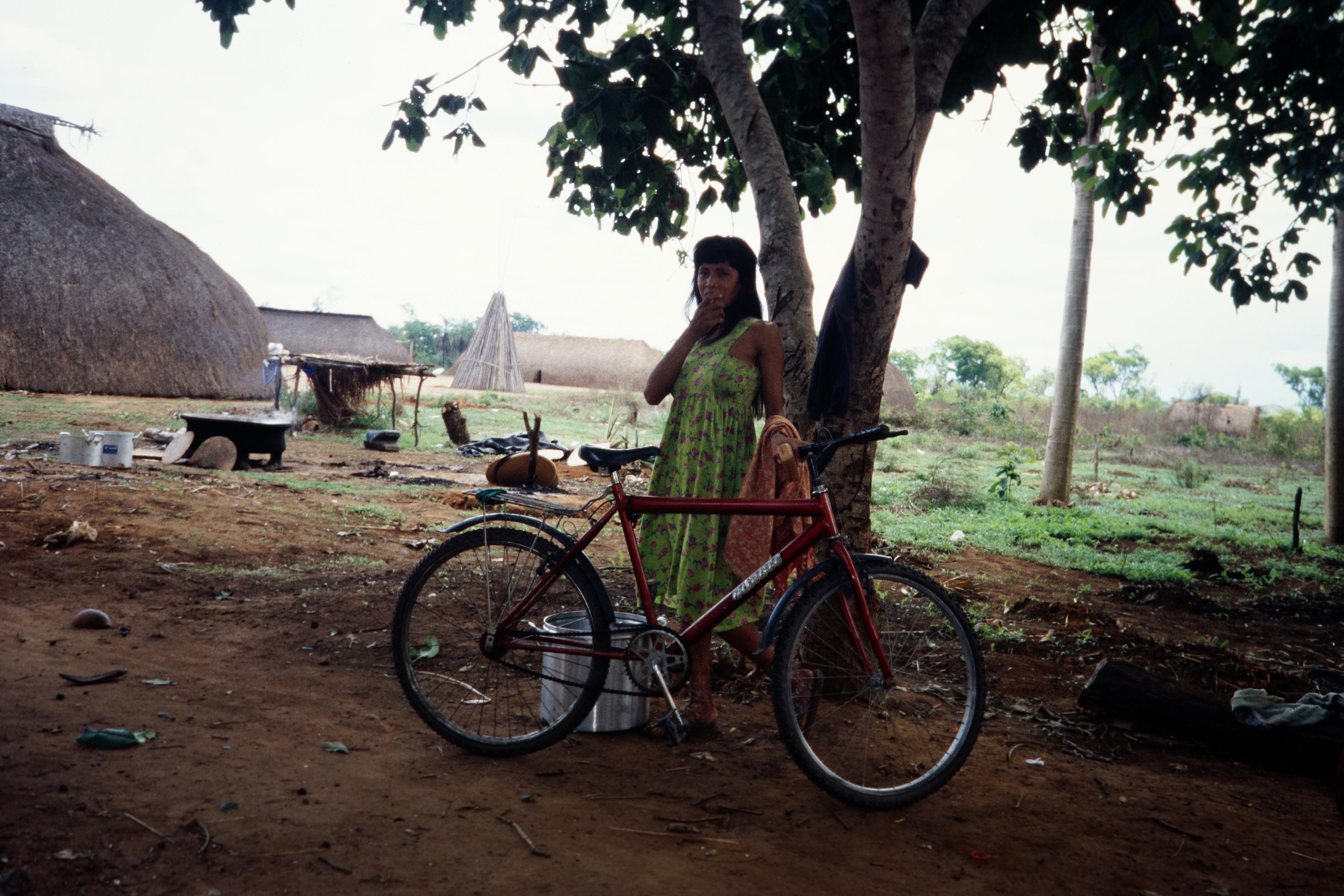 Eine indigene Frau in Brasilien mit einem Metallbehälter und einem Fahrrad im Schatten unter einem Baum.