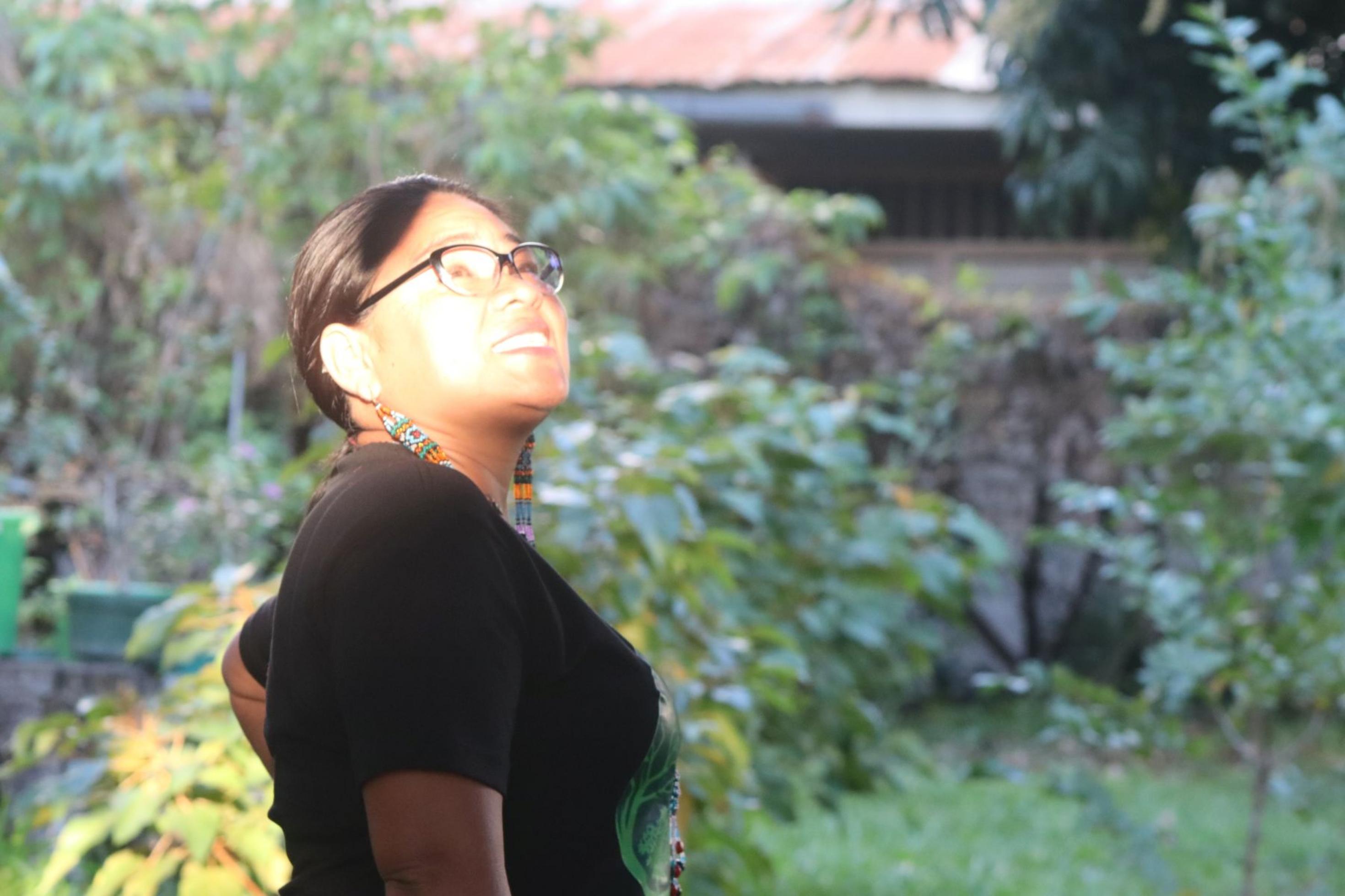 Junge Frau mit nach hinten gebundenen schwarzen Haaren, Brille, blickt schräg rechts nach oben. Die Sonne fällt auf ihre Gesicht. Daneben und im Hintergrund Laubbäume-