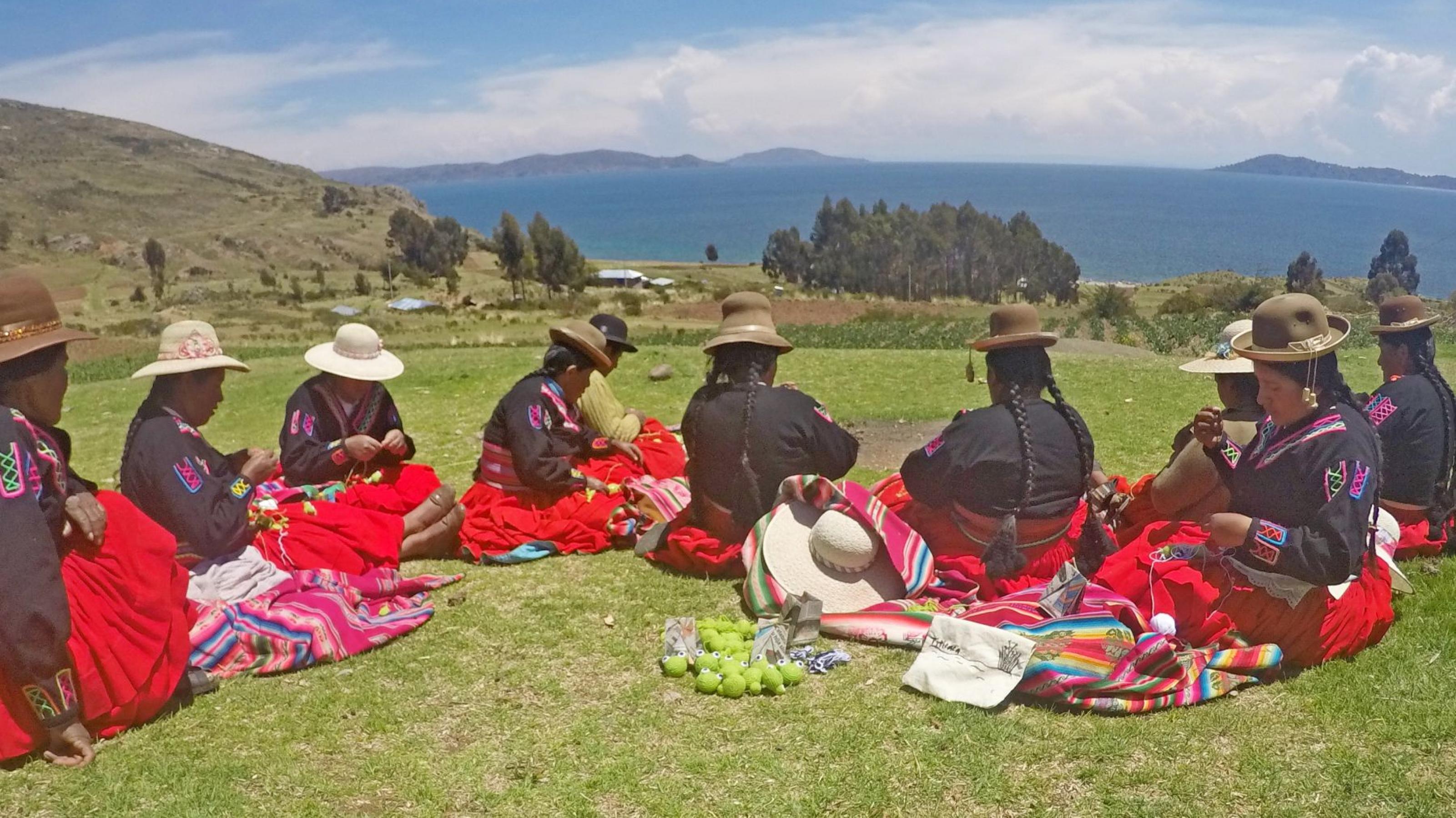 Acht indigene Frauen mit Hut, rotem weiten Röcken und schwarzen Hemden, sitzen auf einem Feld mit Blick auf einen grossen See und  stricken.