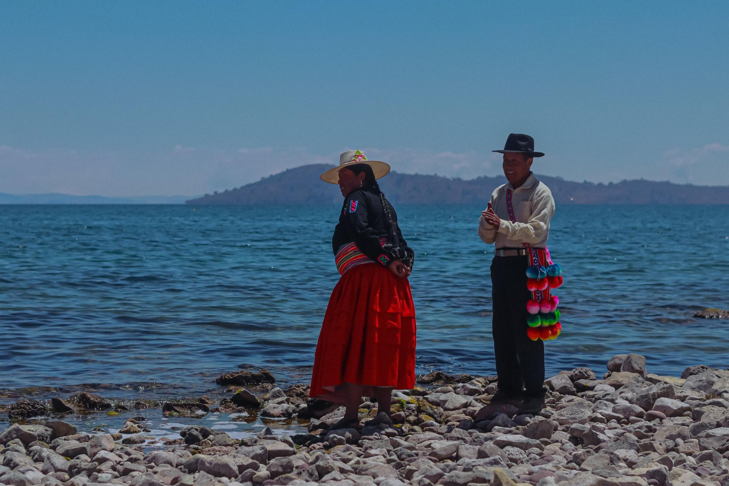 Rückansicht von zwei Menschen, die auf einen grossen See blicken. Sie stehen auf einem Kieselstrand, direkt vor dem Wasser. Es sind zwei indigene Menschen, eine Fraumit weitem rotem Rock und Strohhut und ein Mann mit Hut, weissem Hemd und einer gewebten Hängetasche mit Wollbommeln.