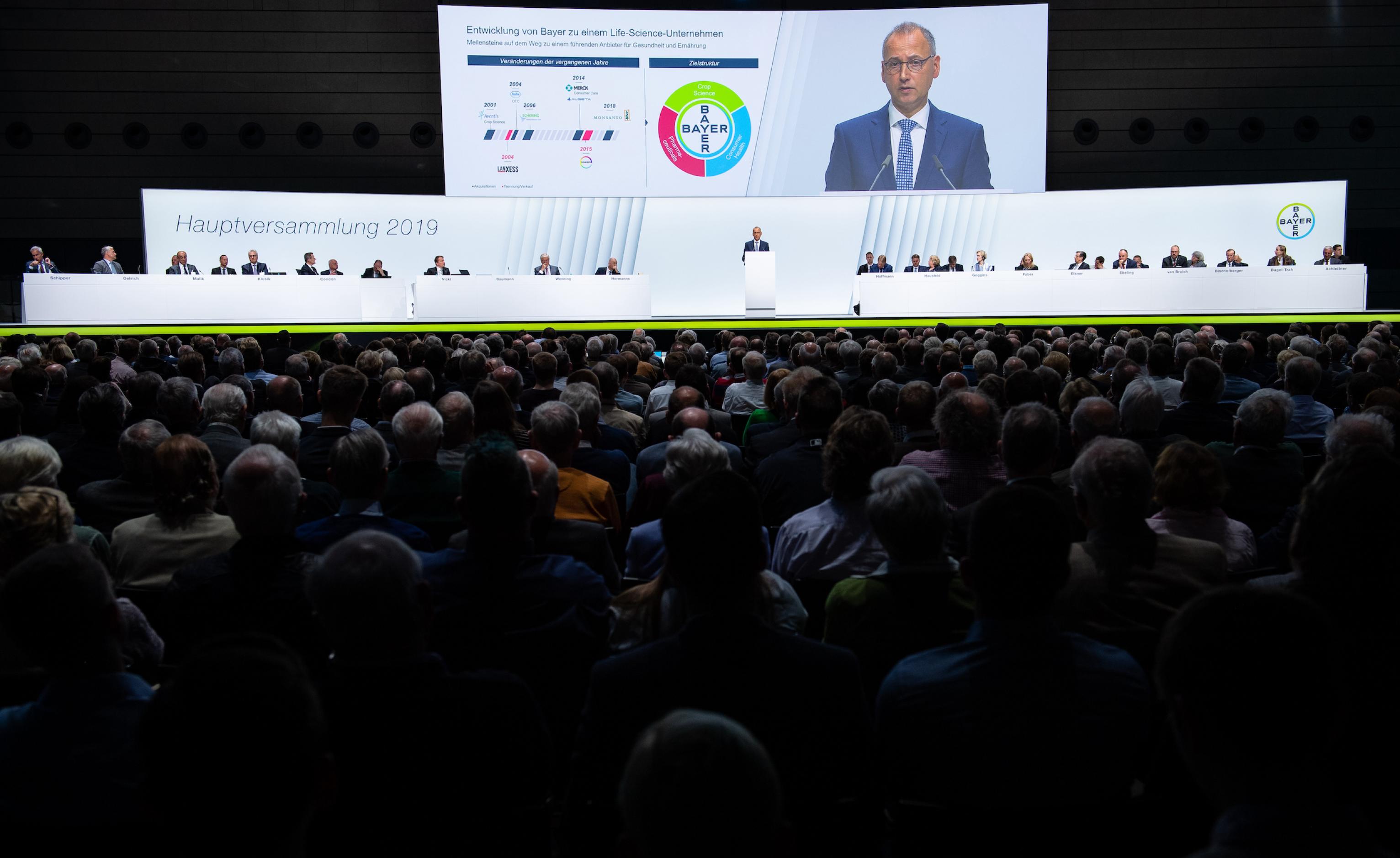 Dunkle Menge sitzender Menschen im Vordergrund, dahinter Podium mit dem Vorstand und Großbildleinwald mit Baumann.