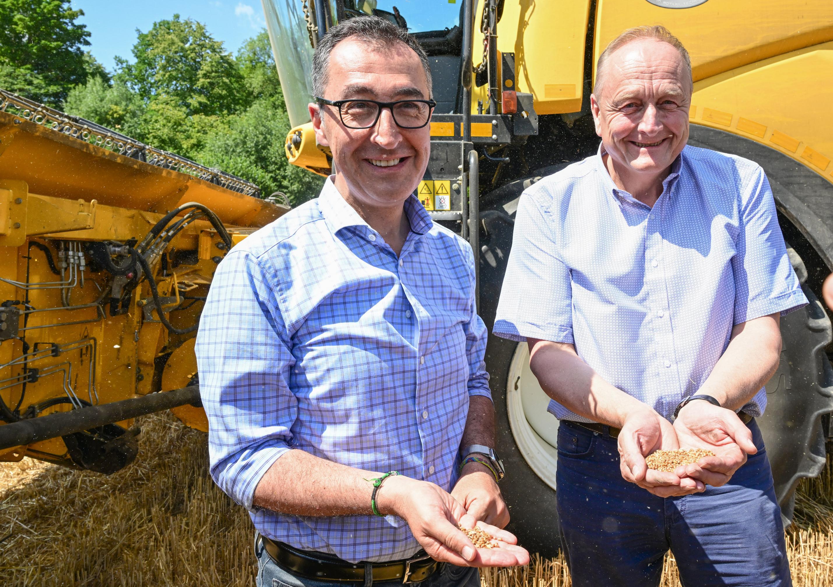 Die beiden Männer stehen vor Agrarmaschinen und halten Getreide in den Händen.