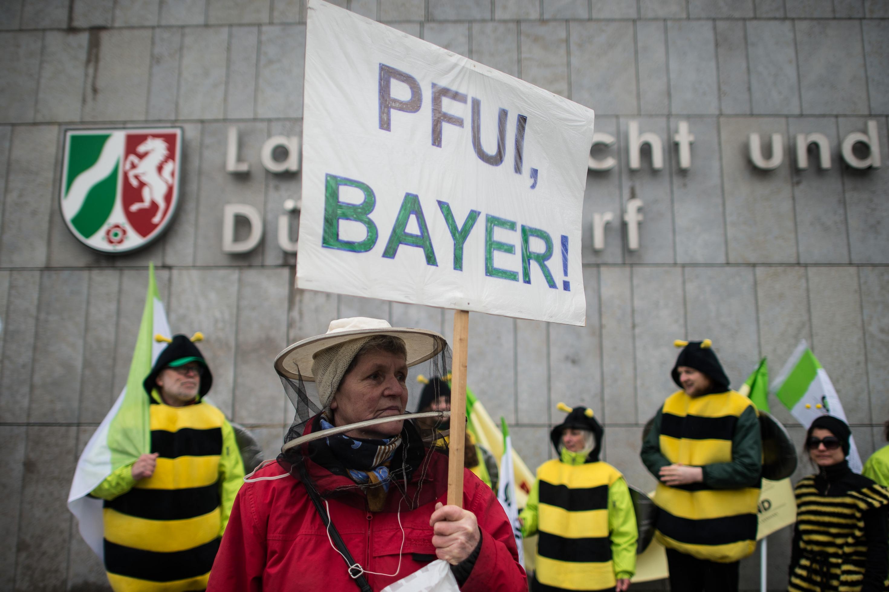 „Pfui, Bayer“ steht auf dem Schild einer Demonstrantin mit Imker-Kopfbedeckung. Hinter ihr drei Menschen in Bienen-Kostümen. Sie stehen vor einem Gerichtsgebäude.