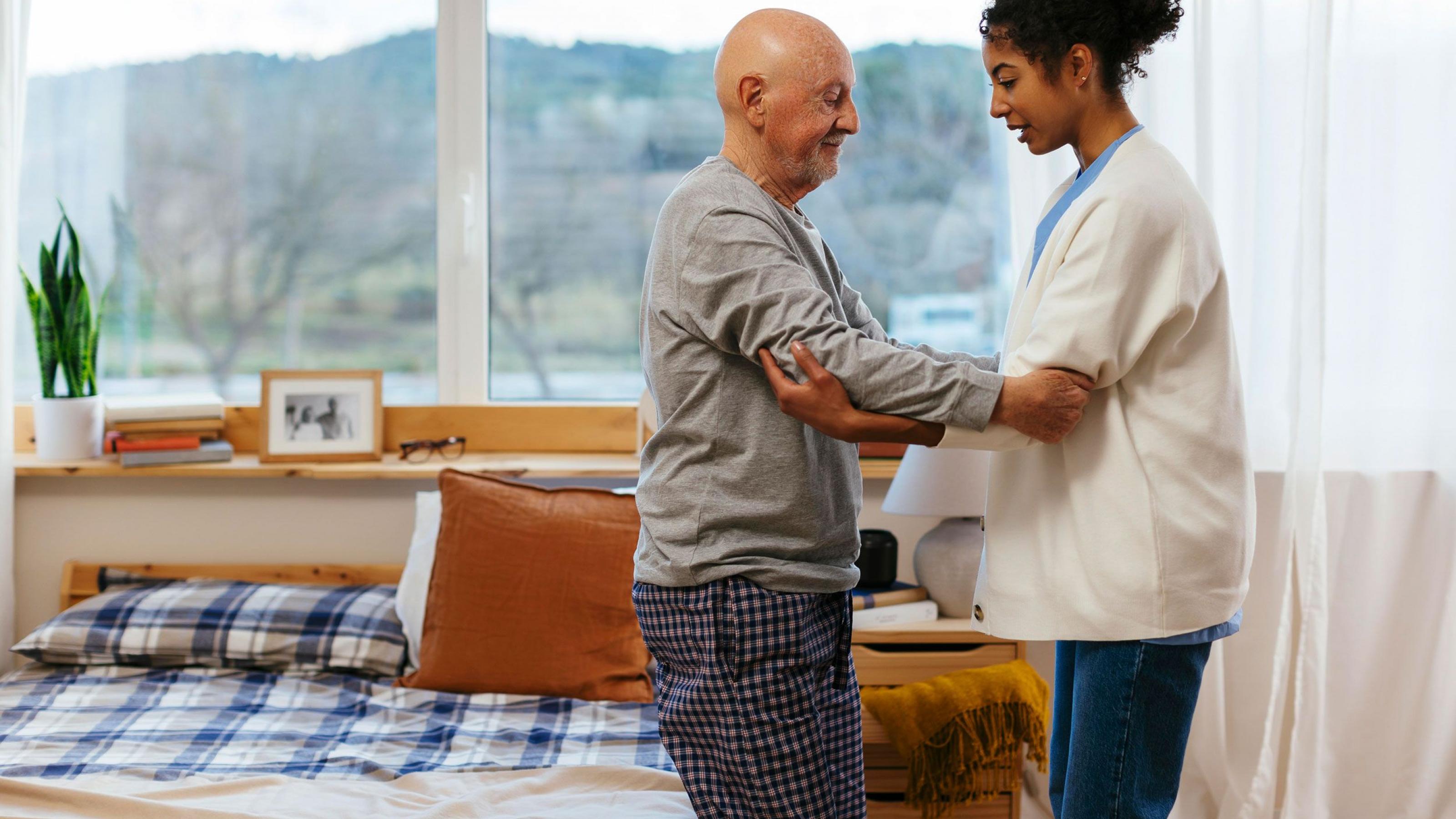 Eine Pflegerin hilft einem alten Mann beim Aufstehen aus seinem Bett. Pflegefachkräfte sind wichtig, um eine häusliche Pflege zu unterstützen.