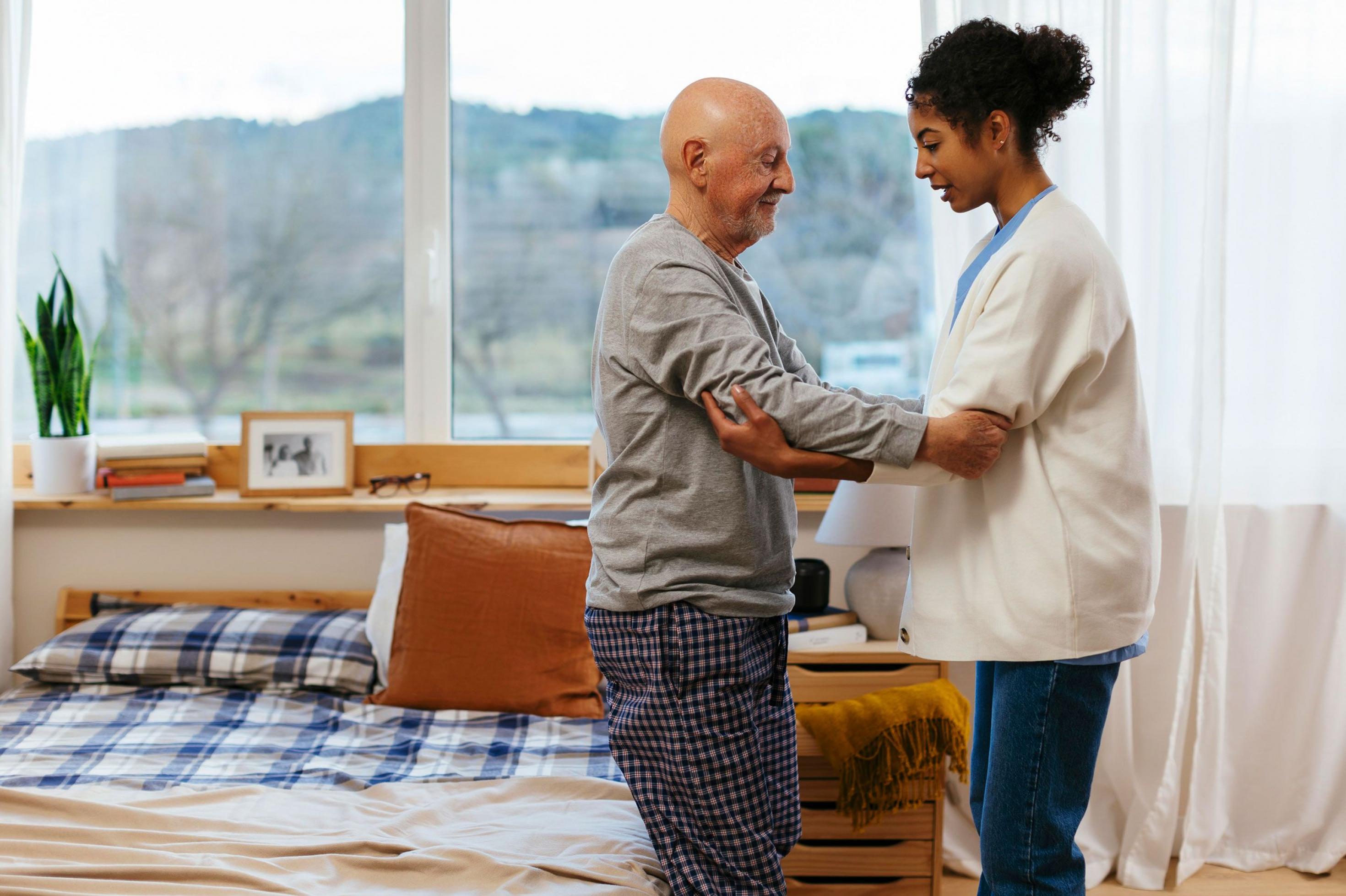 Eine Pflegerin hilft einem alten Mann beim Aufstehen aus seinem Bett. Pflegefachkräfte sind wichtig, um eine häusliche Pflege zu unterstützen.
