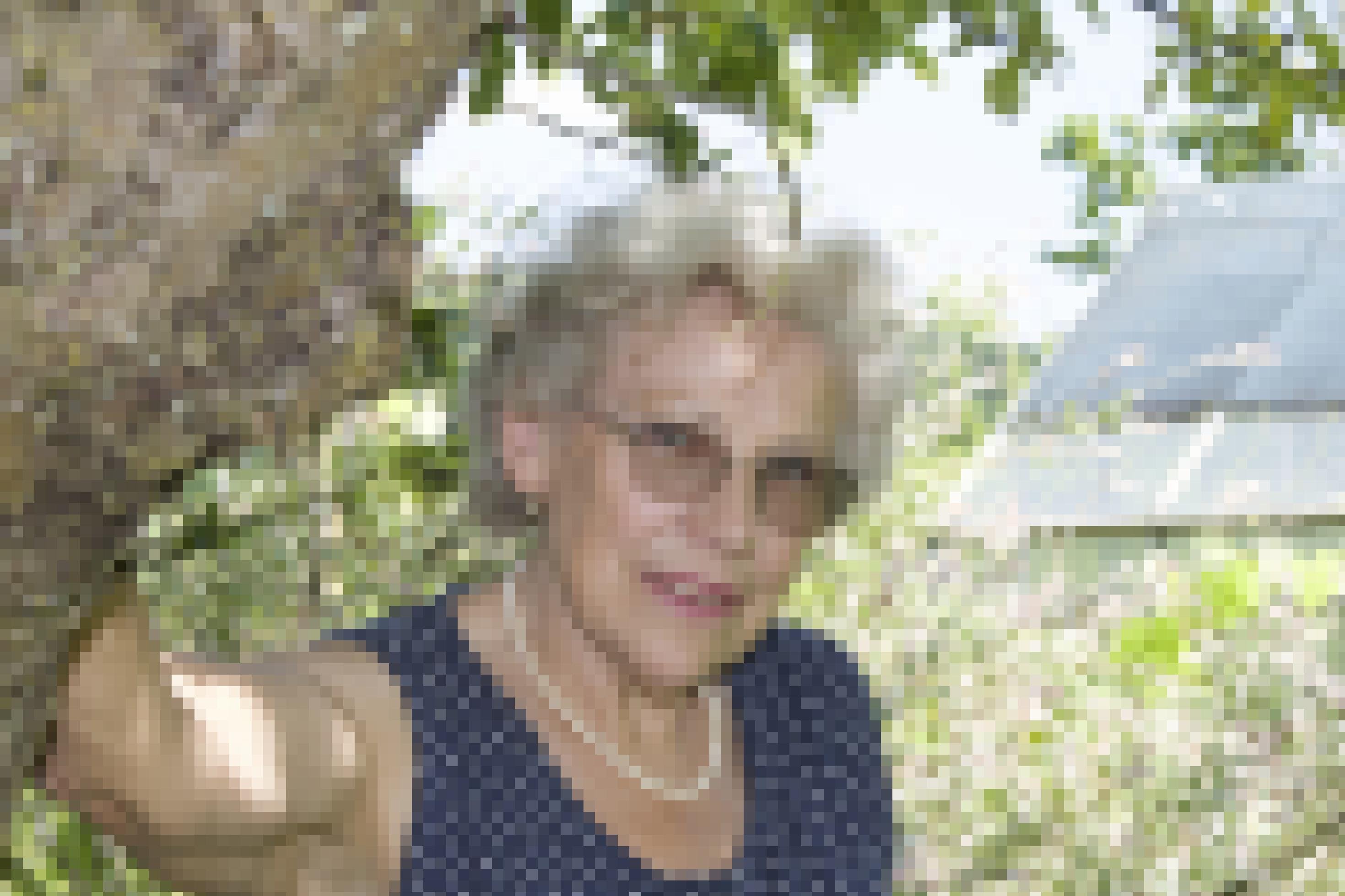 Eine ältere Frau mit kurzen grauweißen Locken, randloser, Brille, Perlenkette und einem ärmellosen blauen Shirt mit weißen Punkten steht in einem Garten und lehnt sich mit der Hand leicht an einen Baumstamm.