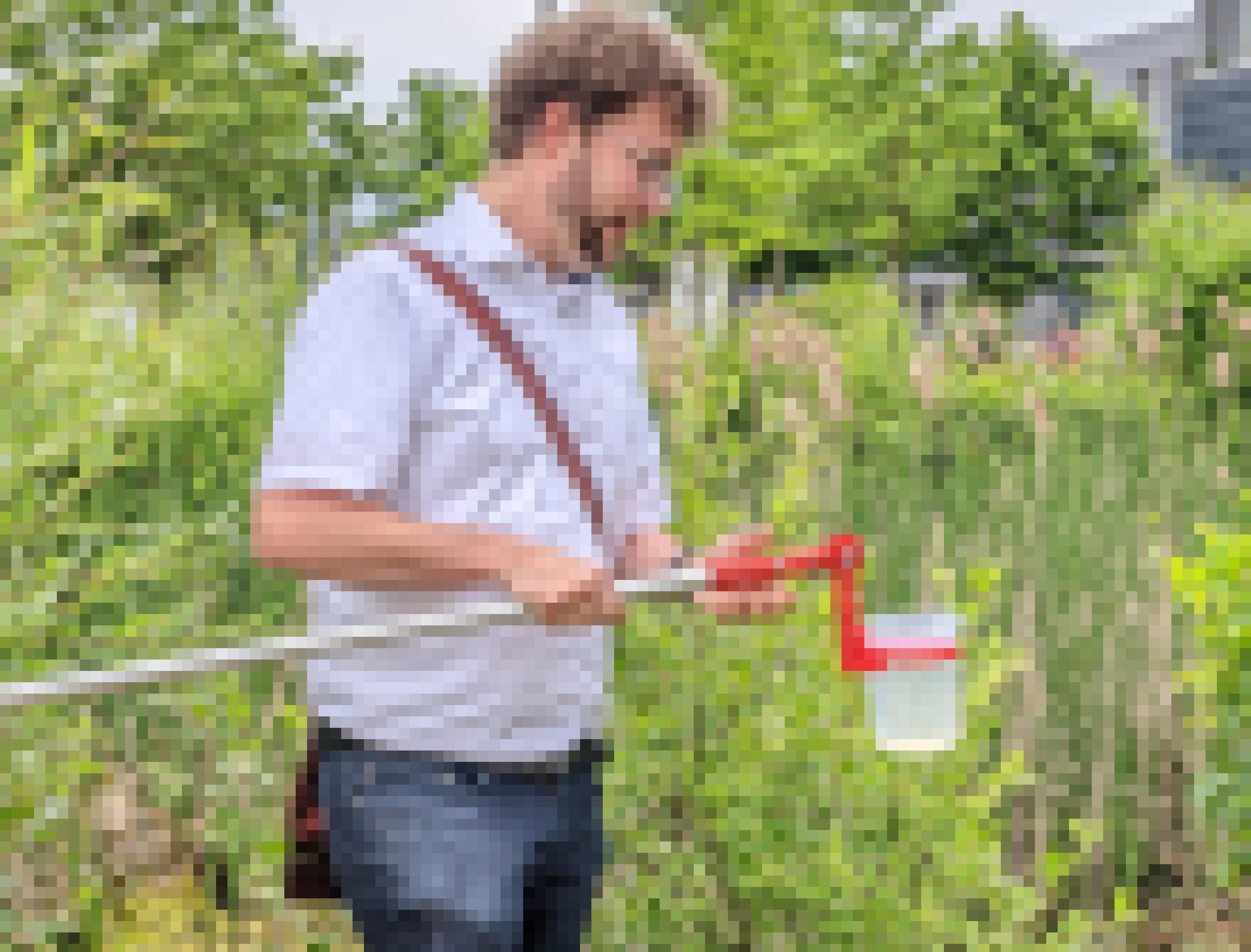 Ein Mann in Jeans und Kurzarm-Hemd steht zwischen Schilf und Büschen. Er hat eine lange Stange in der Hand. Vorne an der Stange ist ein großer Plastikbecher  befestigt, in dem ein bisschen Wasser zu sehen ist.