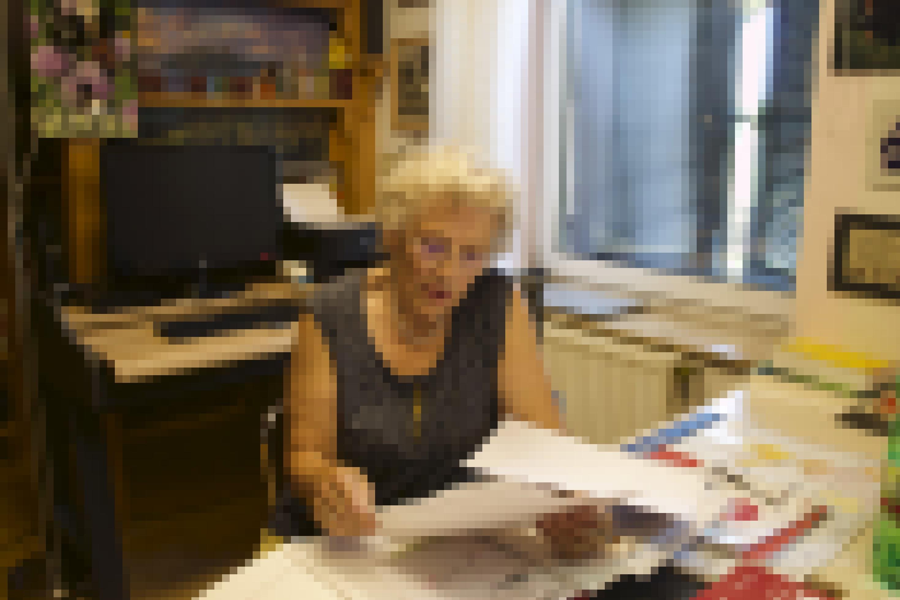 Eine ältere Frau mit kurzen weiß-grauen Locken, Brille und Perlenkette sitzt an einem Schreibtisch. Sie hält einen dünnen Stapel A4-Papiere in der Hand und blickt auf eines davon. Im Hintergrund sind ein Computerbildschirm und eine Tastatus sowie ein Fenster mit fast geschlossenen Fensterläden zu sehen.