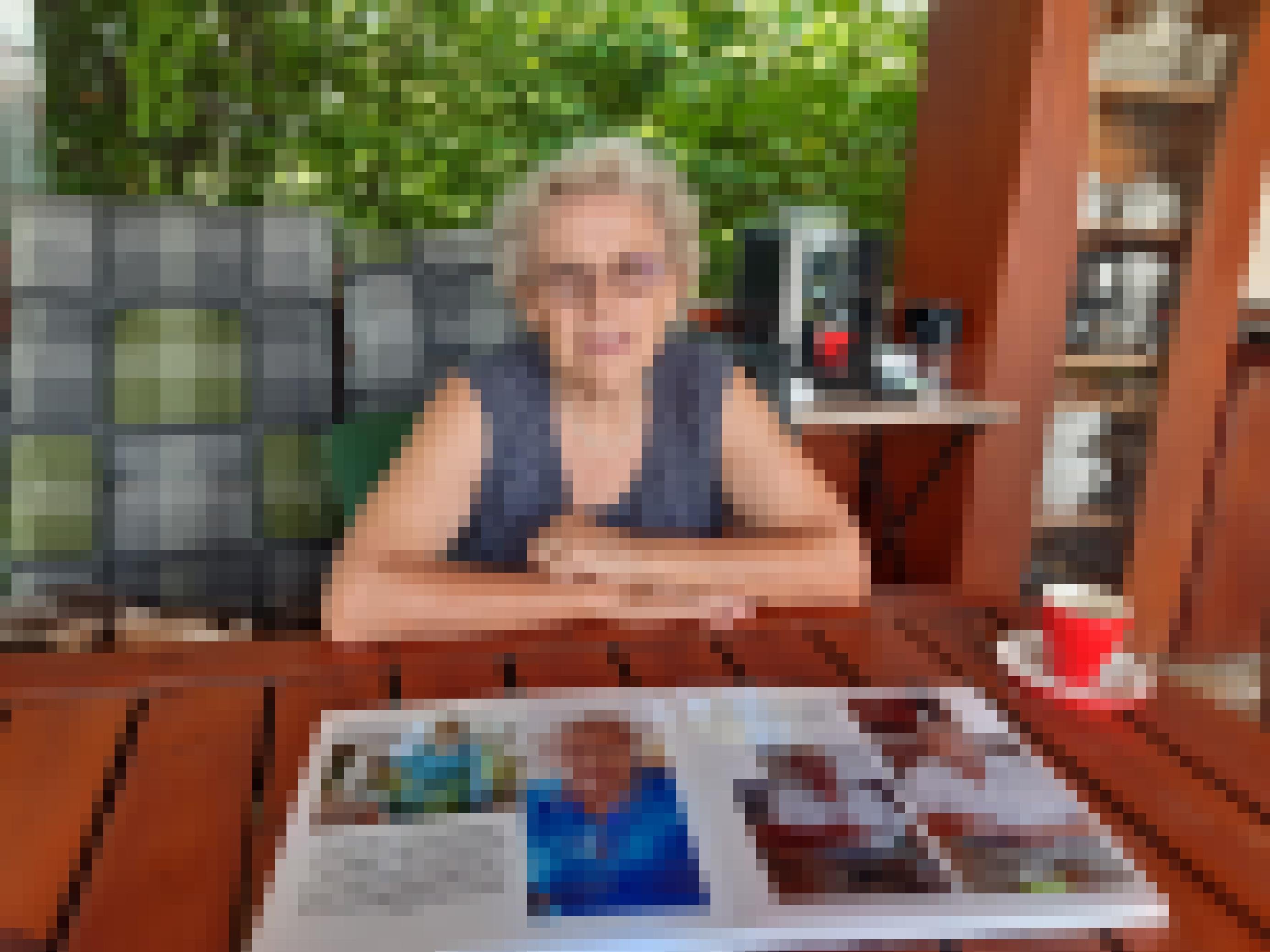 Eine ältere Frau mit kurzen grau-weißen Locken, Brille und Perlenkette sitzt auf einer gepolsterten Bank in einem Garten. Auf dem Holztisch vor ihr liegt ein aufgeschlagenes Buch. Darin sind je zwei Fotos von ihr und einem Mann erkennbar.