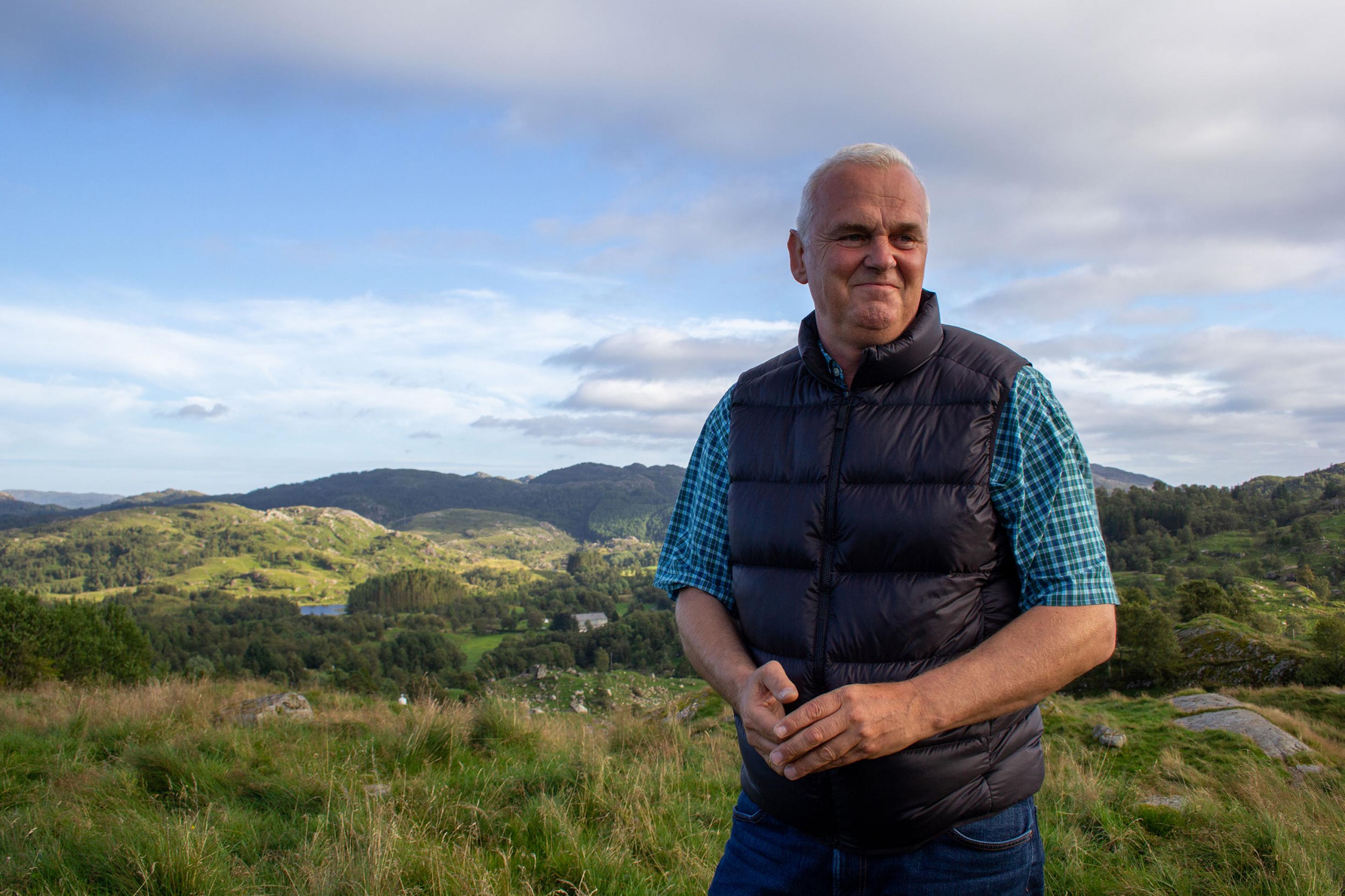 Ein Mann im mittleren Alter, kurze graue Haare, in Daunenweste und kariertem Hemd in einer weiten, hügeligen Landschaft