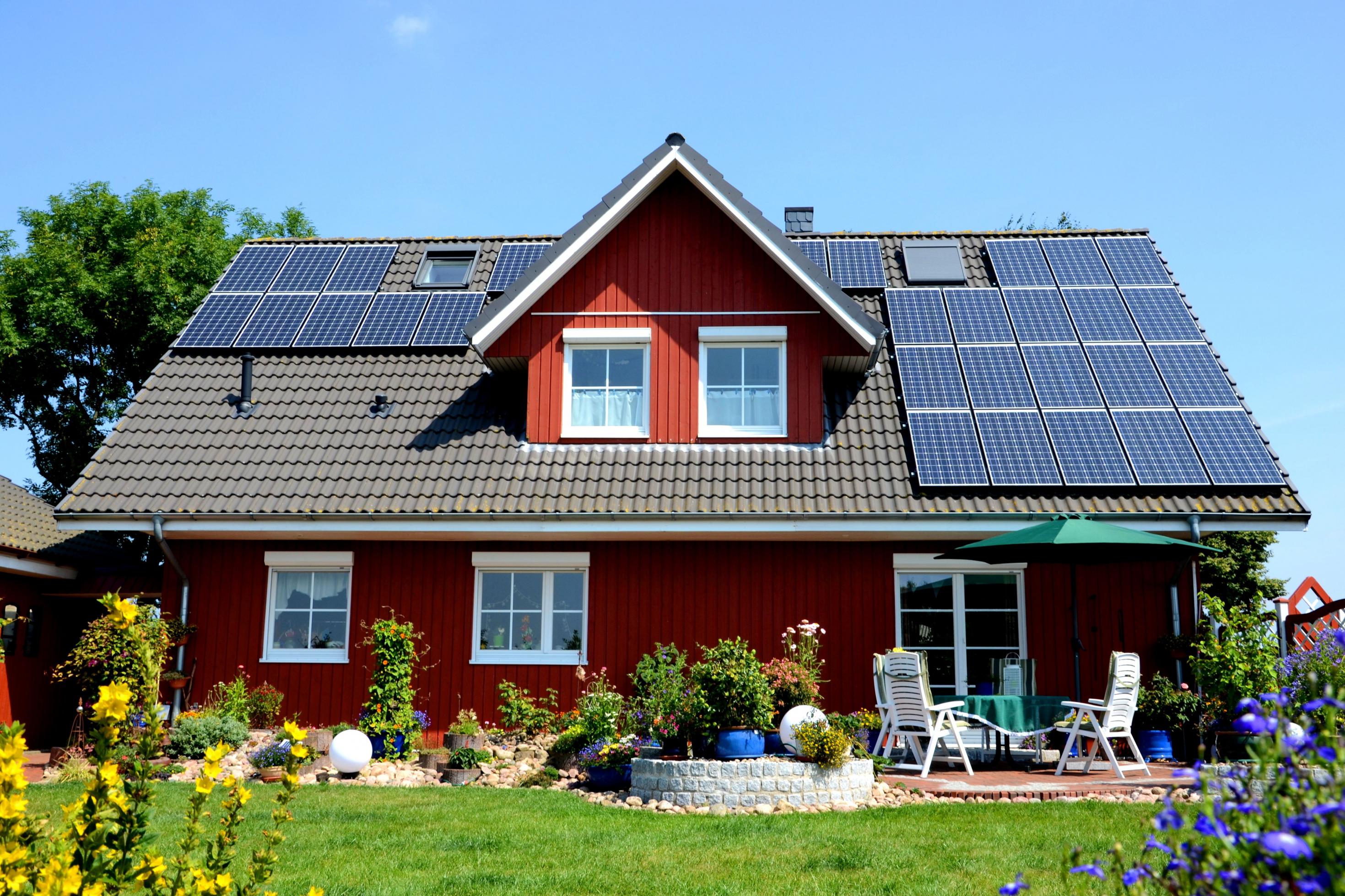 Frontalansicht eines roten Einfamilienhauses mit großer Photovoltaik-Anlage auf dem Dach, im Vordergrund Garten