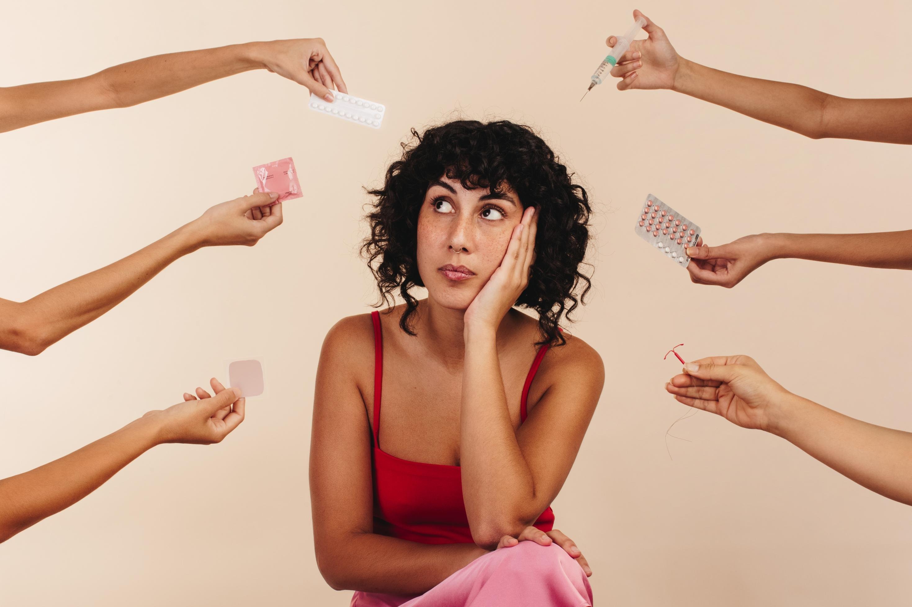 Eine Frau vor einem rosa Hintergrund betrachtet nachdenklich verschiedene Verhütungsmittel.