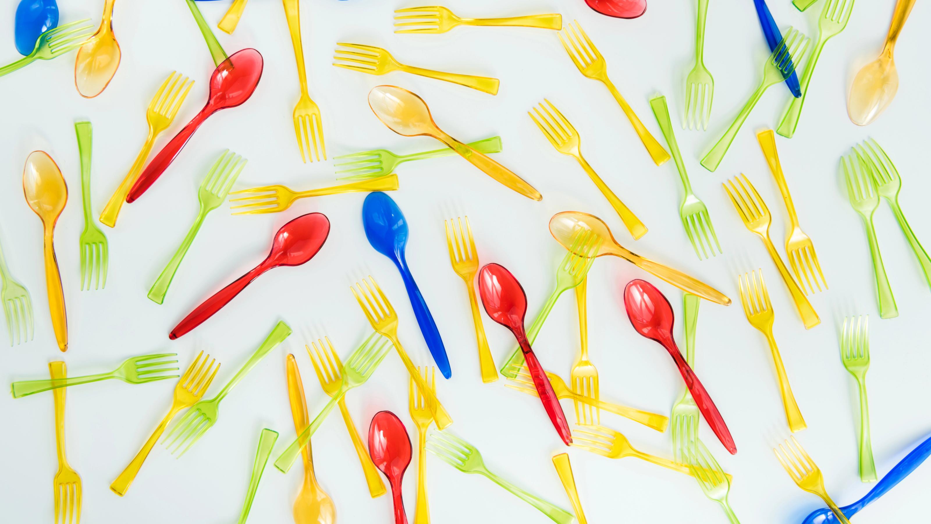 Blaue, gelbe, grüne und rote Plastik-Gabeln und Plastik-Löffel, durcheinander liegend auf einem weißen Untergrund.