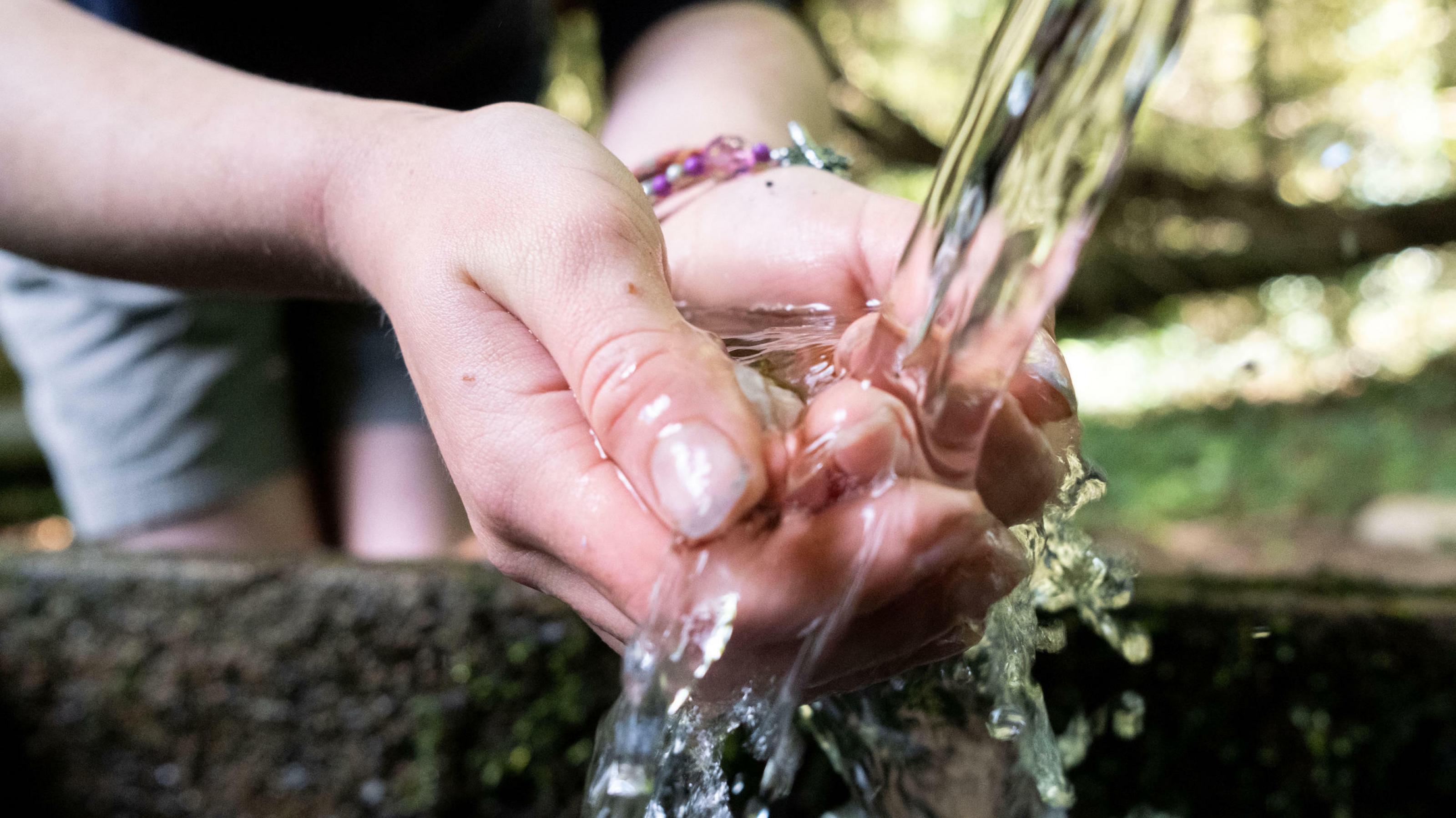 Eine junge Frau schöpft mit den Händen frisches Wasser aus dem Strahl eines Brunnens.