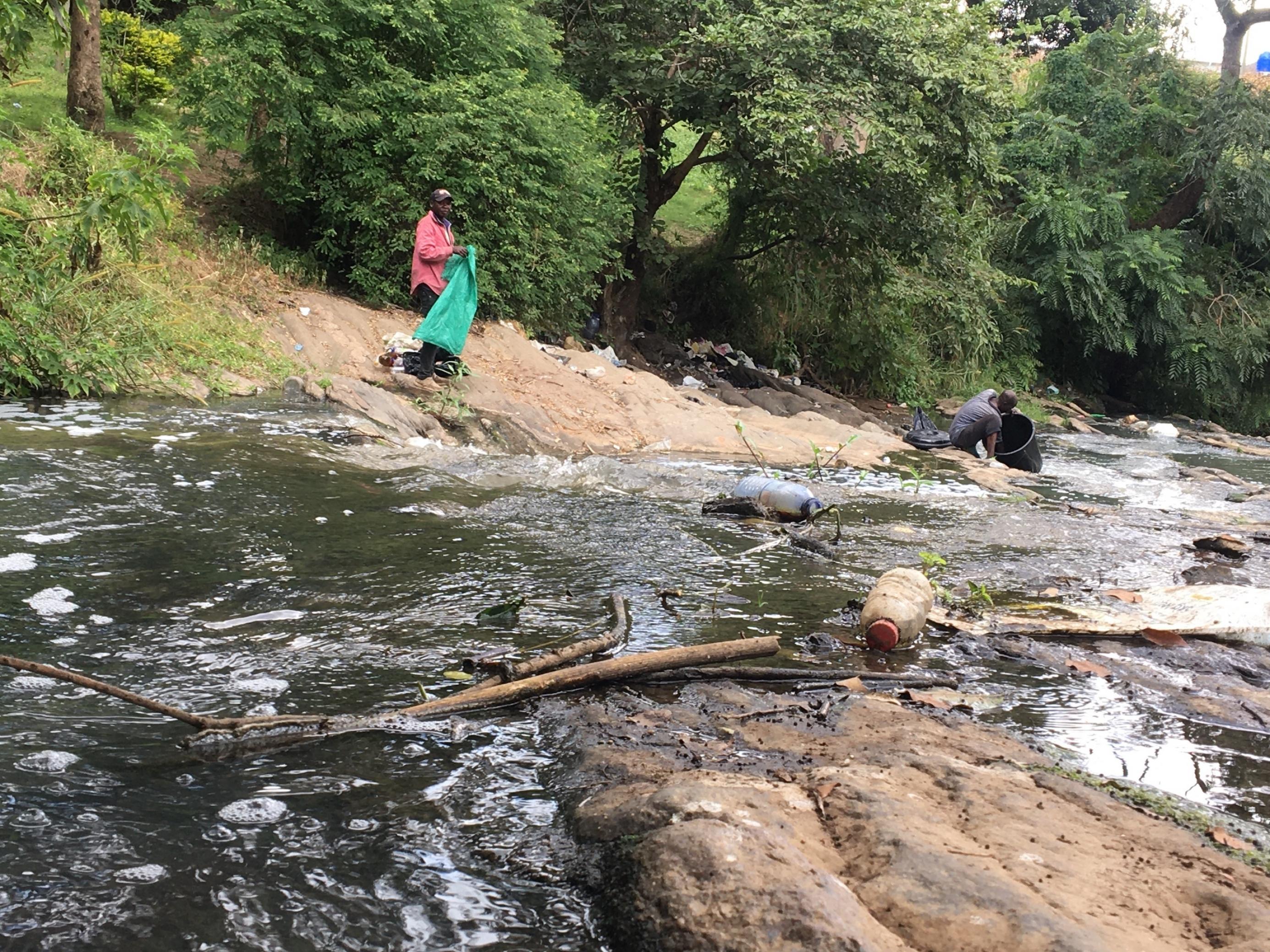 Zwei Männer an einem mit Plastikmüll verschmutzten Fluss in Malawi. Einer hat eine Mülltüte in der Hand, der andere sammelt den Abfall ein.