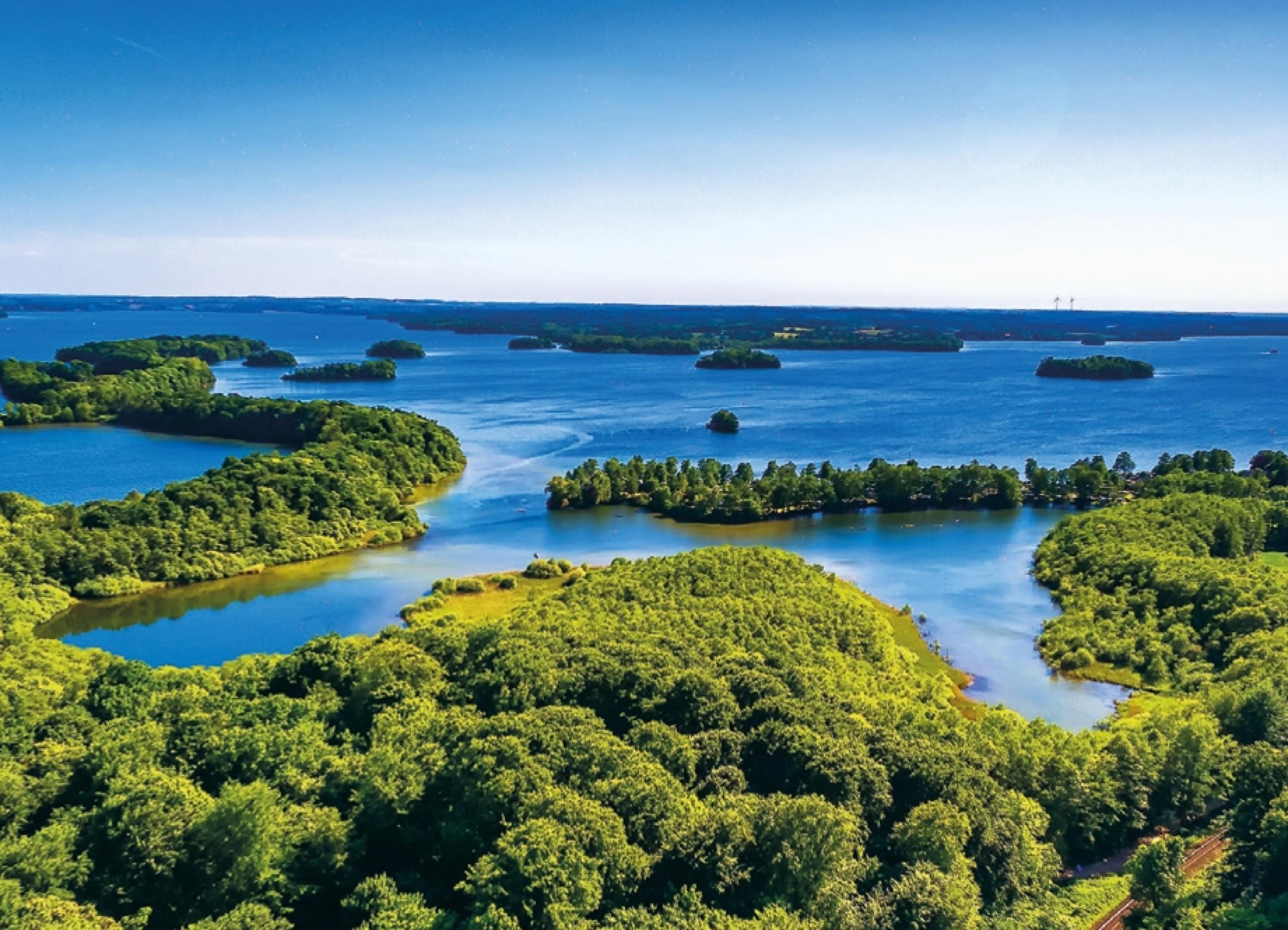 Unter blauem Himmel erstrecken sich zahlreiche tiefblaue Wasserflächen, die von baumreichem Grün umrandet werden.