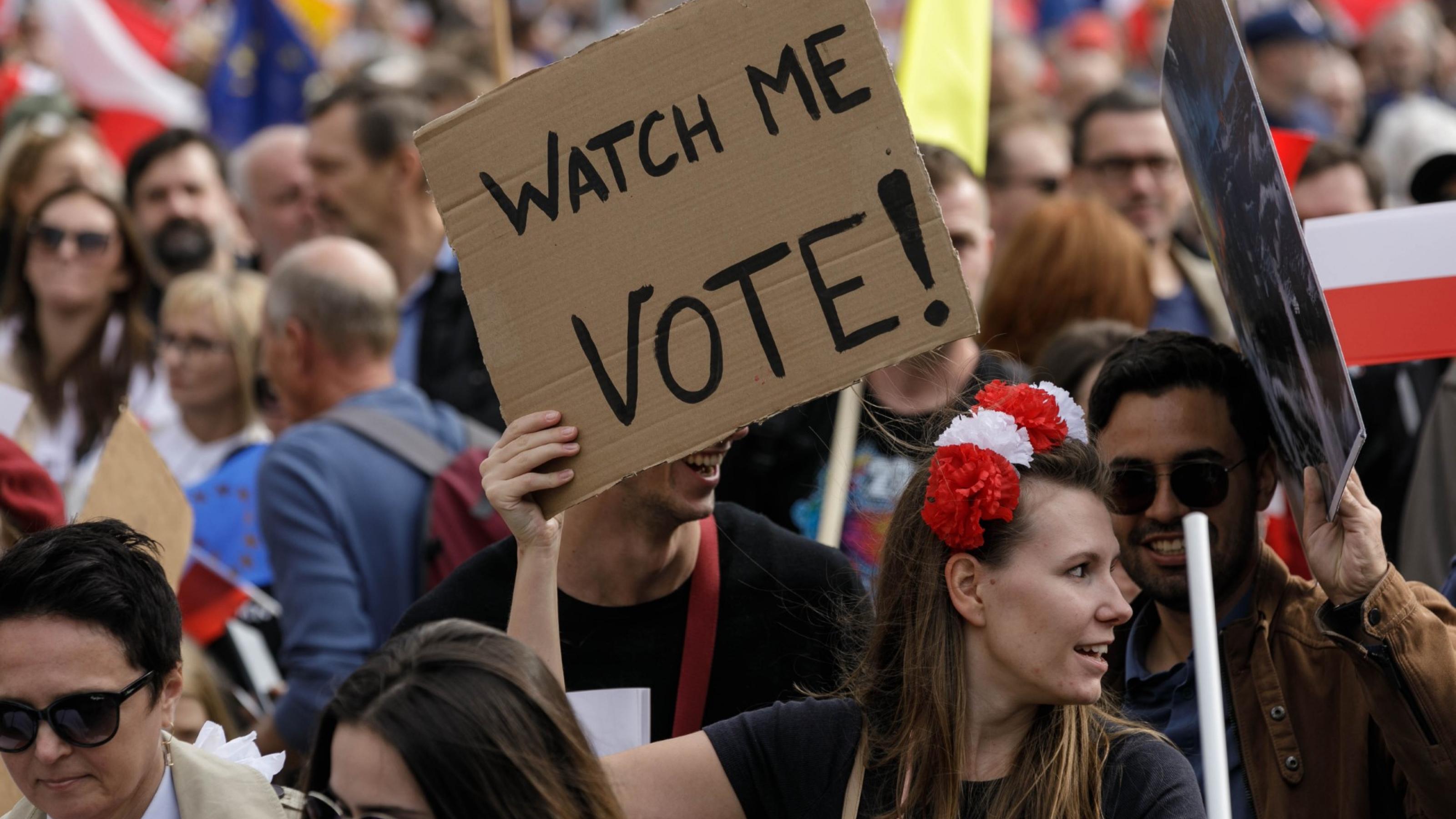 eine Gruppe von Menschen, die Schilder halten. Eine Frau hält ein Schild mit dem Spruch „Watch me Vote“