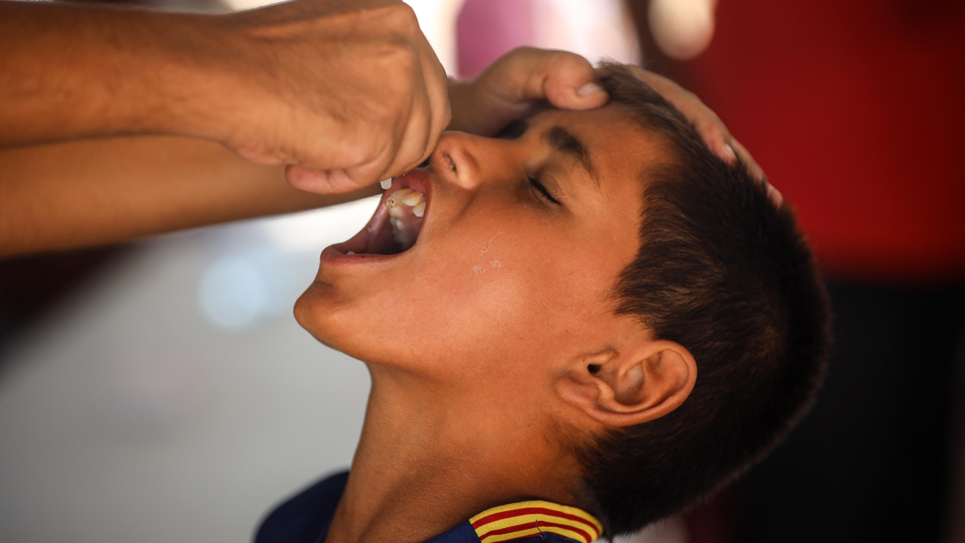 Ein Junge mit dunklen kurzen Haaren, steht mit geöffnetem Mund vor einer anderen Person, die ihm aus einer kleinen Plastikampulle eine Impflösung in den Mund tropft.