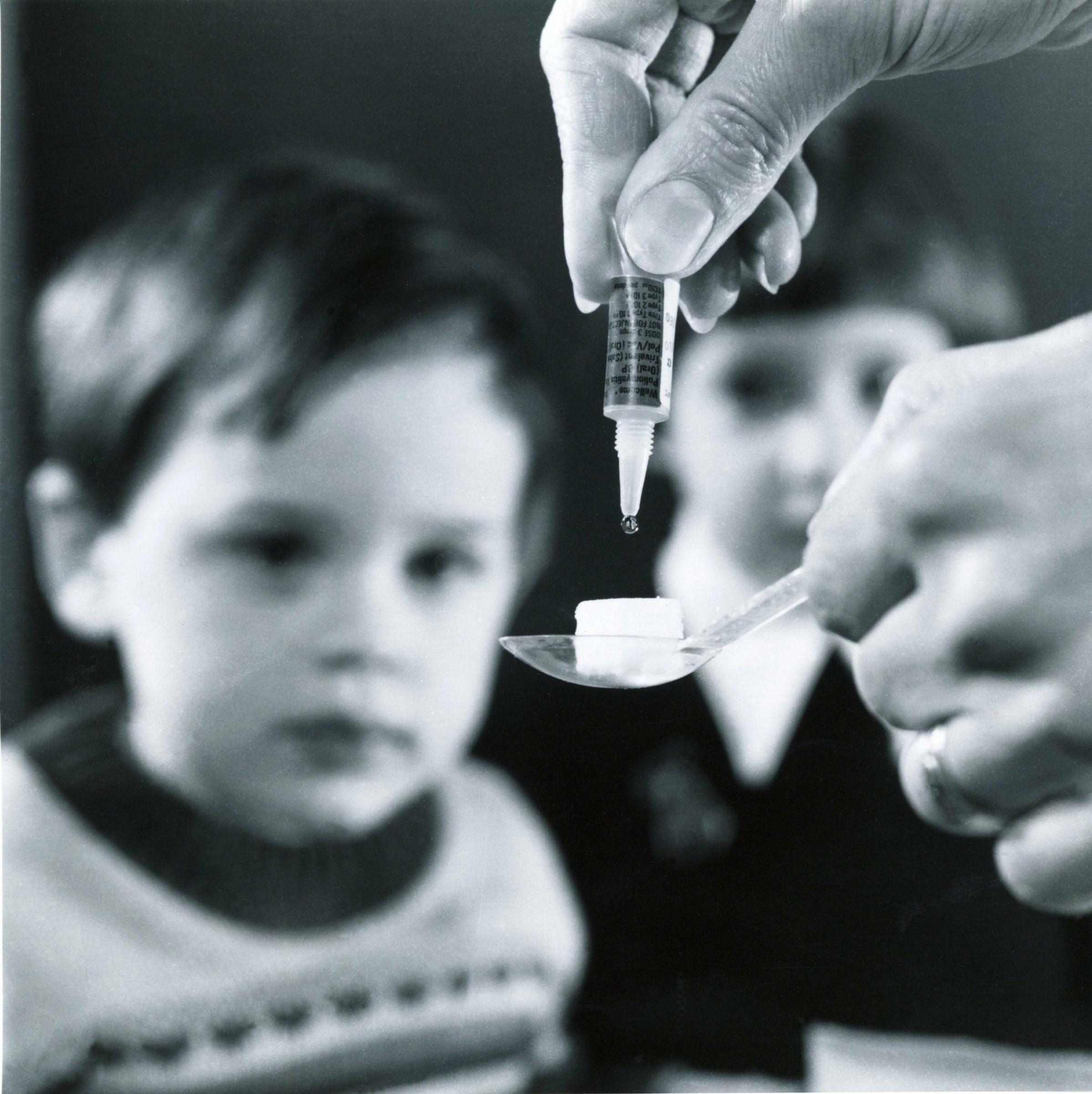 Zwei Kinder schauen auf diesem Schwarz-Weiß-Foto zu, wie eine ärztliche Fachkraft den Polioimpfstoff auf ein Zuckerstückchen träufelt.