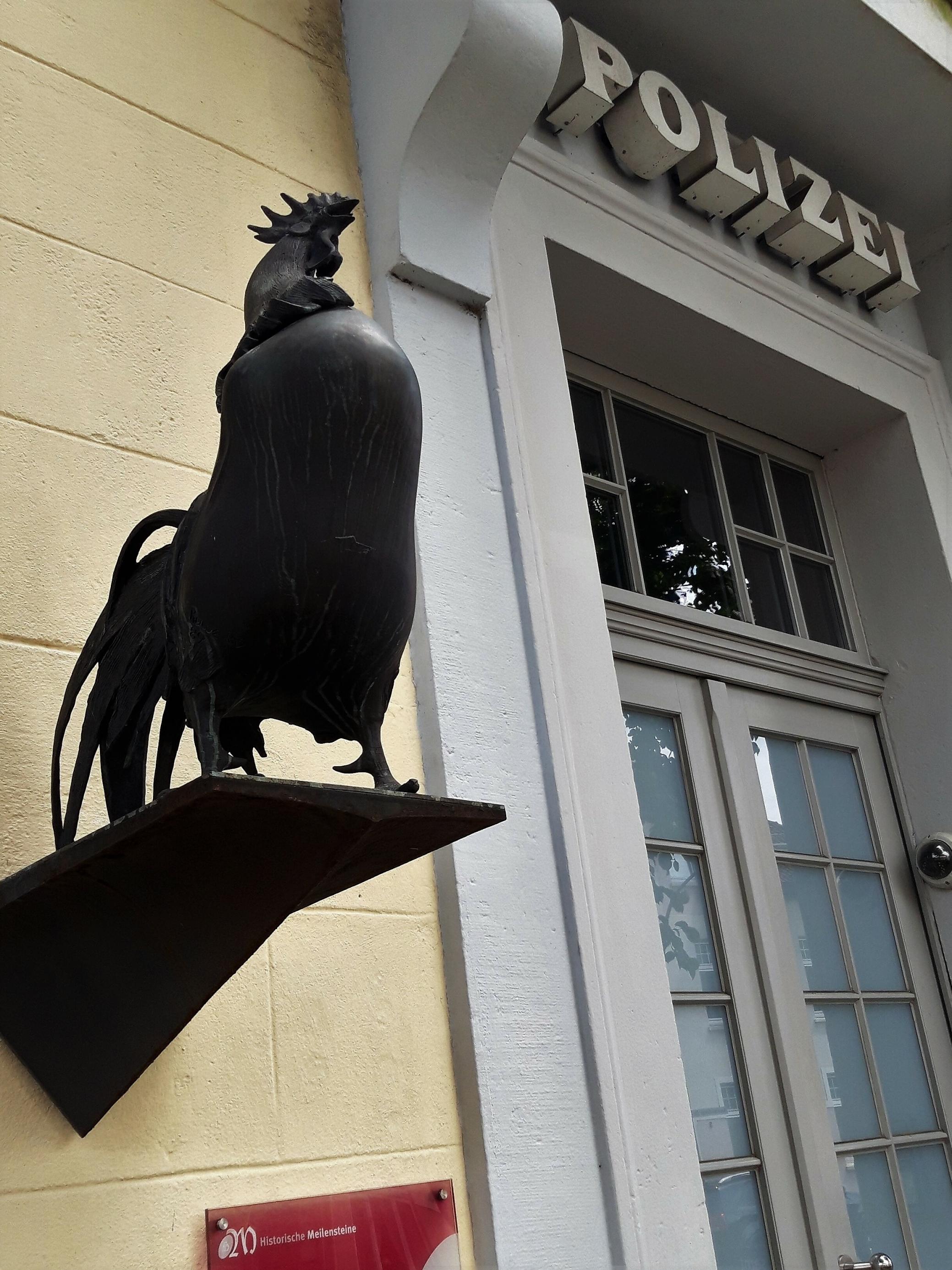 Im Vordergrund ist die an der Außenwand angebrachte Bronzeplastik zu sehen, im Hintergrund die Eingangstür, überschrieben mit „Polizei“.