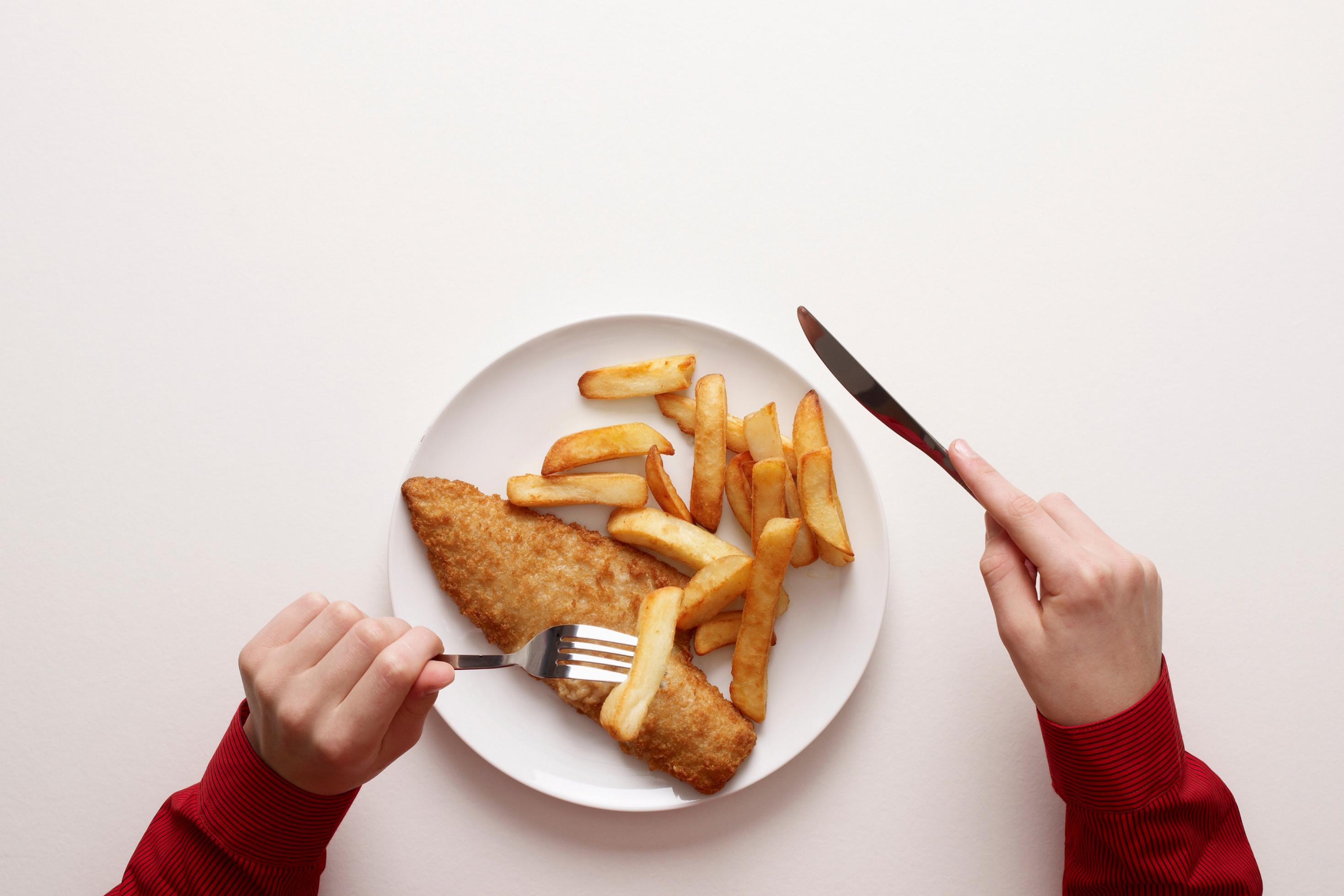 Draufsicht: In der Mitte steht ein weißer Teller, darauf etliche Pommes und ein Stück paniertes Fleisch. Kinderhände halten Messer und Gabel.
