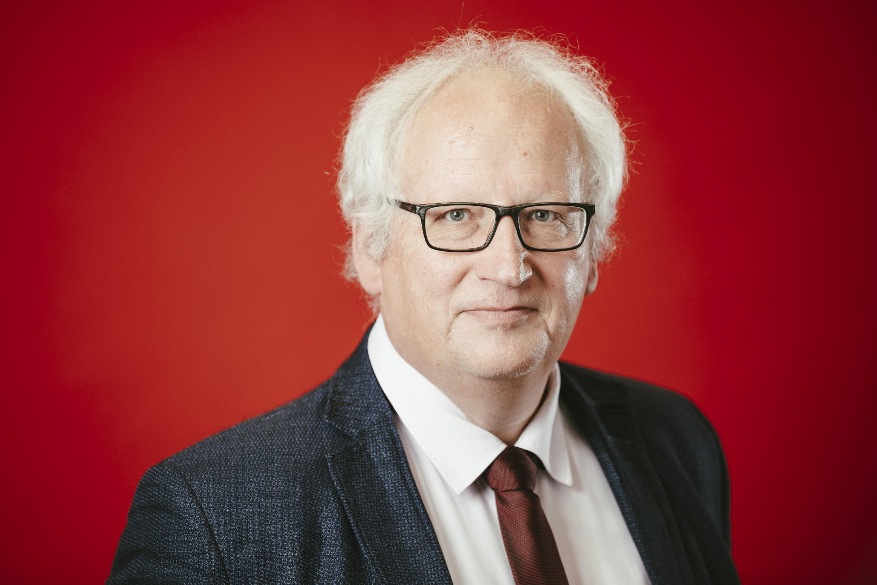 Bernhard Emunds von der Hochschule Sankt Georgen in Frankfurt/Main hat weiße Haare und trägt eine dunkle Brille. Das Portrait zeigt in in Anzug und Krawatte vor rotem Hintergrund.