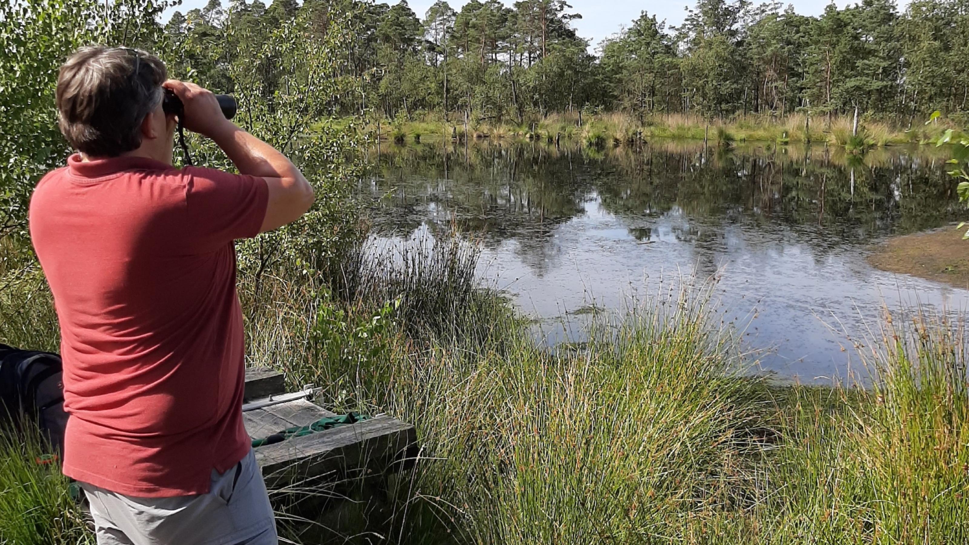 Eick von Ruschkowski steht vor einem Teich. Er schaut durch ein Fernglas.