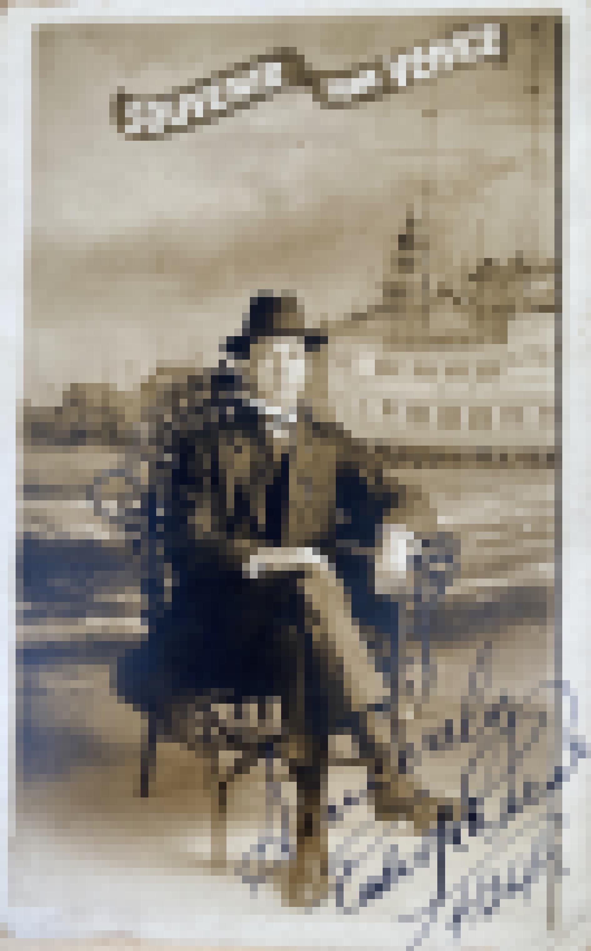 Formelles Portrait von Edward Reese, sitzend in einem Fotostudio. Reese trägt Anzug und Hut und hat die Beine übereinander geschlagen. Hinter ihm eine Fototapete mit Strand und Pier.