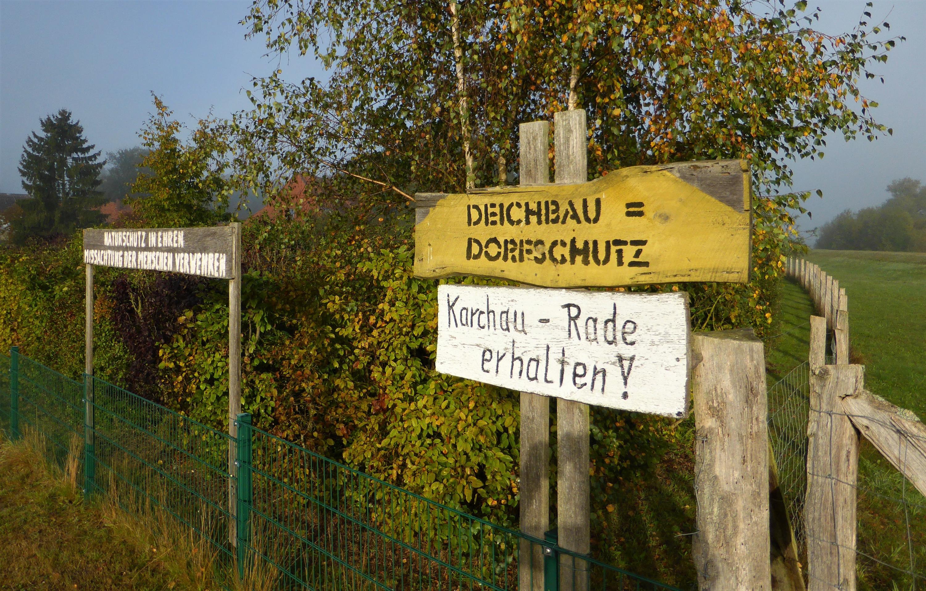 „Deichbau = Dorfschutz“ und „Naturschutz in Ehren – Missachtung der Menschen verwehren“: Dies Inschriften stehen auf Holzlatten am Rand eines Grundstückzauns, hinter dem rechts ein Deichabschnitt zu sehen ist.