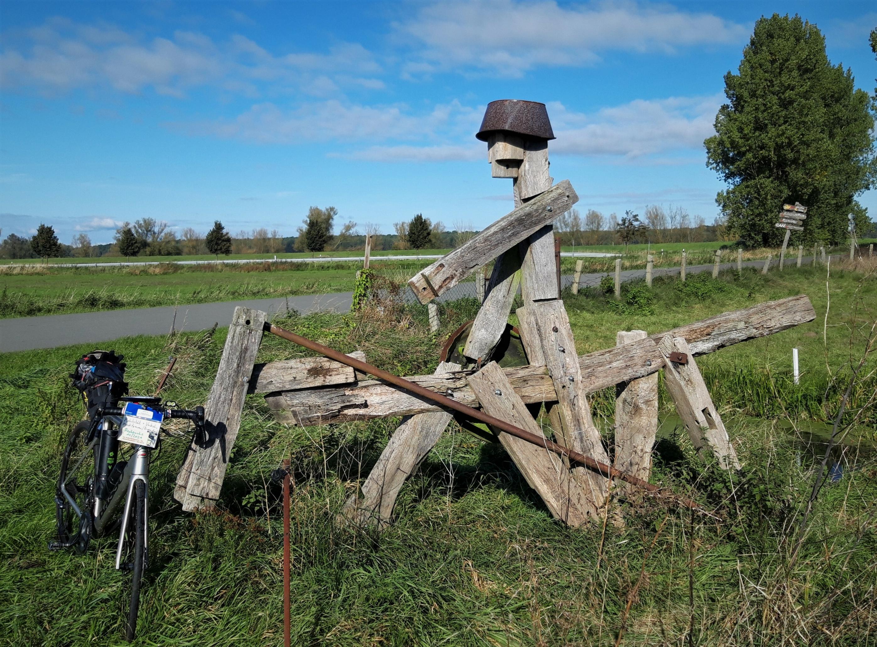 Stilisiertes verwittertes Holzmonument eines Bleicheimer-behelmten Reiters, dessen Metall-Lanze vom ausgestreckten Arm gefallen ist. Am Kopf des Holzpferdes lehnt das Rennrad des Reporters. Die Landschaft dahinter ist grün unter blauem Himmel; einziges weiteres Zeichen der Zivilisation ist eine schmale leere Landstraße.