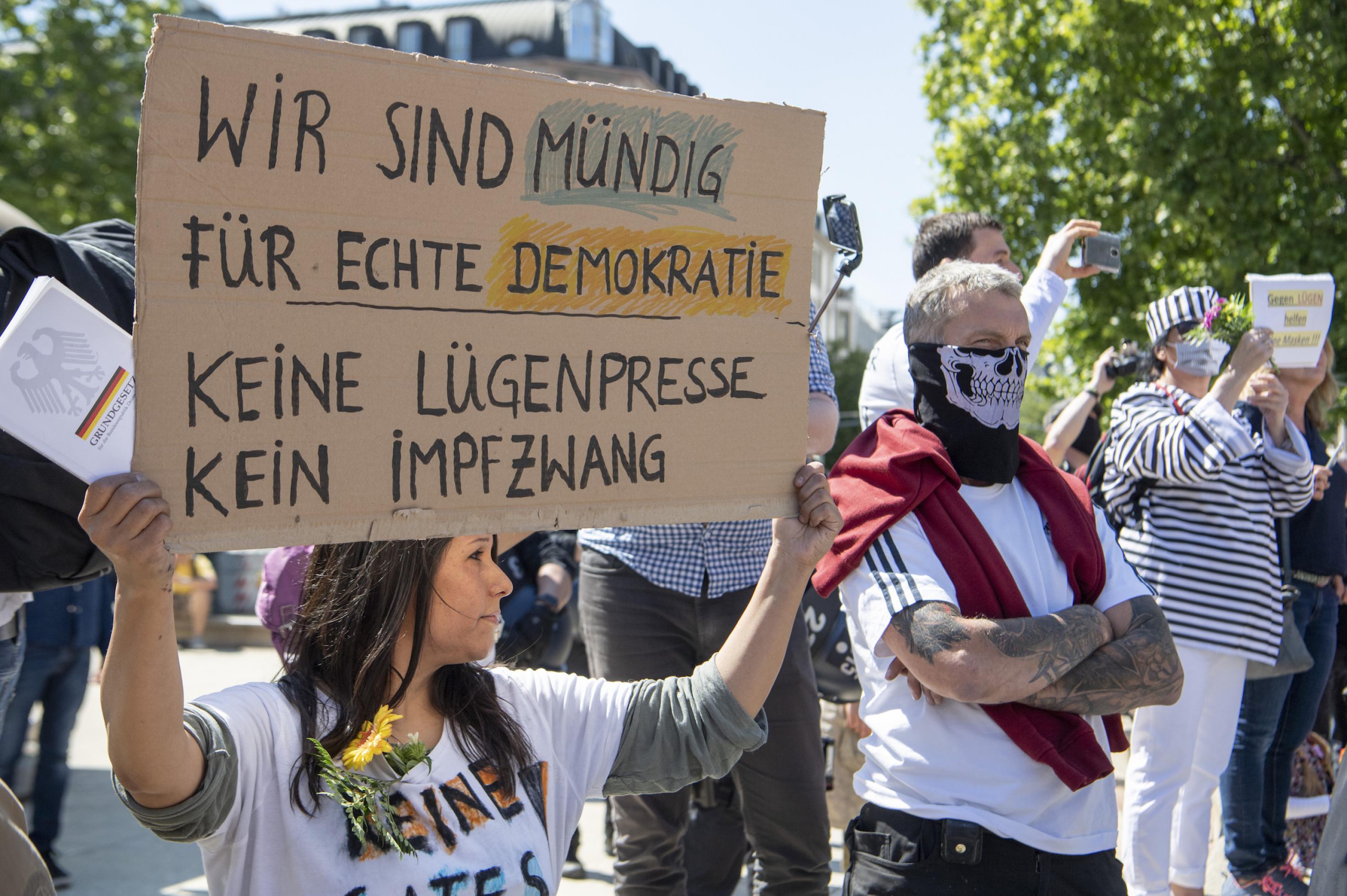 Demonstrantinnen und Demonstranten. Eine Frau hält ein Pappschild hoch, auf dem steht: „Wir sind mündig für echte Demokratie – Keine Lügenpresse. Kein Impfzwang“. Neben ihr steht ein Mann, dessen untere Gesichtshälfte mit einem Tuch verdeckt ist, auf dem ein Totenschädel zu sehen ist. Daneben eine Person in Sträflingskleidung.