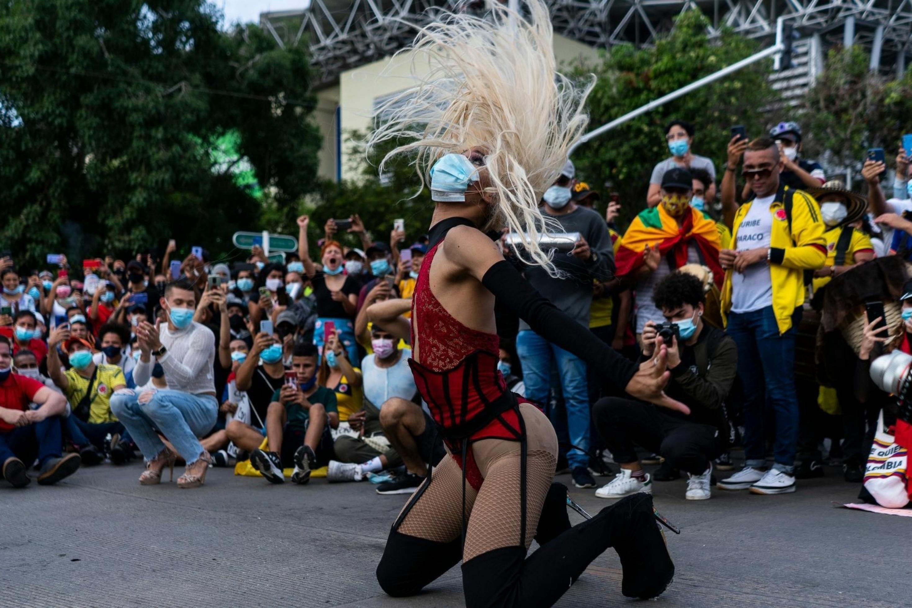 Eine Transfrau mit Mundschutz, hochhackigen Stiefeln und hautengem Spitzenkorsett tanzt auf dem Boden. Sie ist umgeben von  Zuschauerïnnen. Ihre angen Haare stehen vor lauter Schwung senkrecht in der Luft.