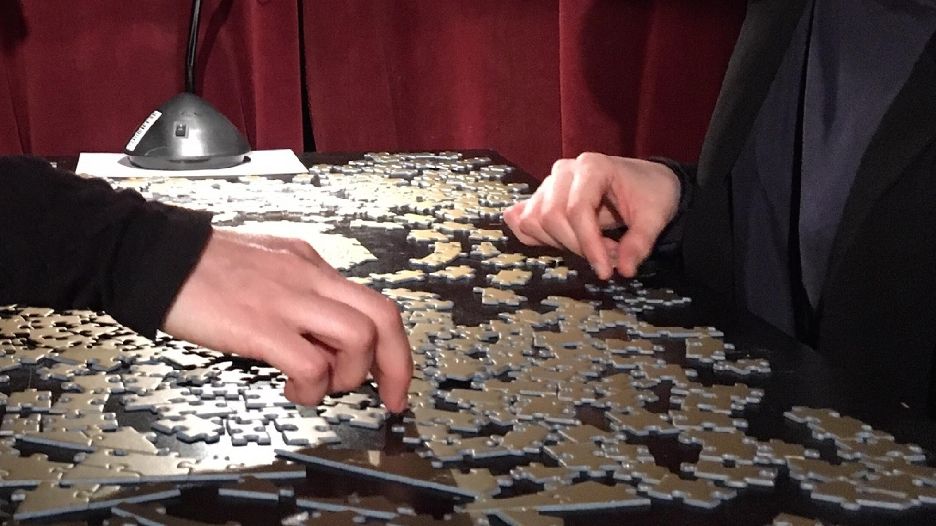 Zwei Menschen sitzen sich gegenüber und puzzlen gemeinsam. Man sieht nur ihre Hände. Die einfarbig goldenen Puzzleteile unterscheiden sich nur durch ihre Form.