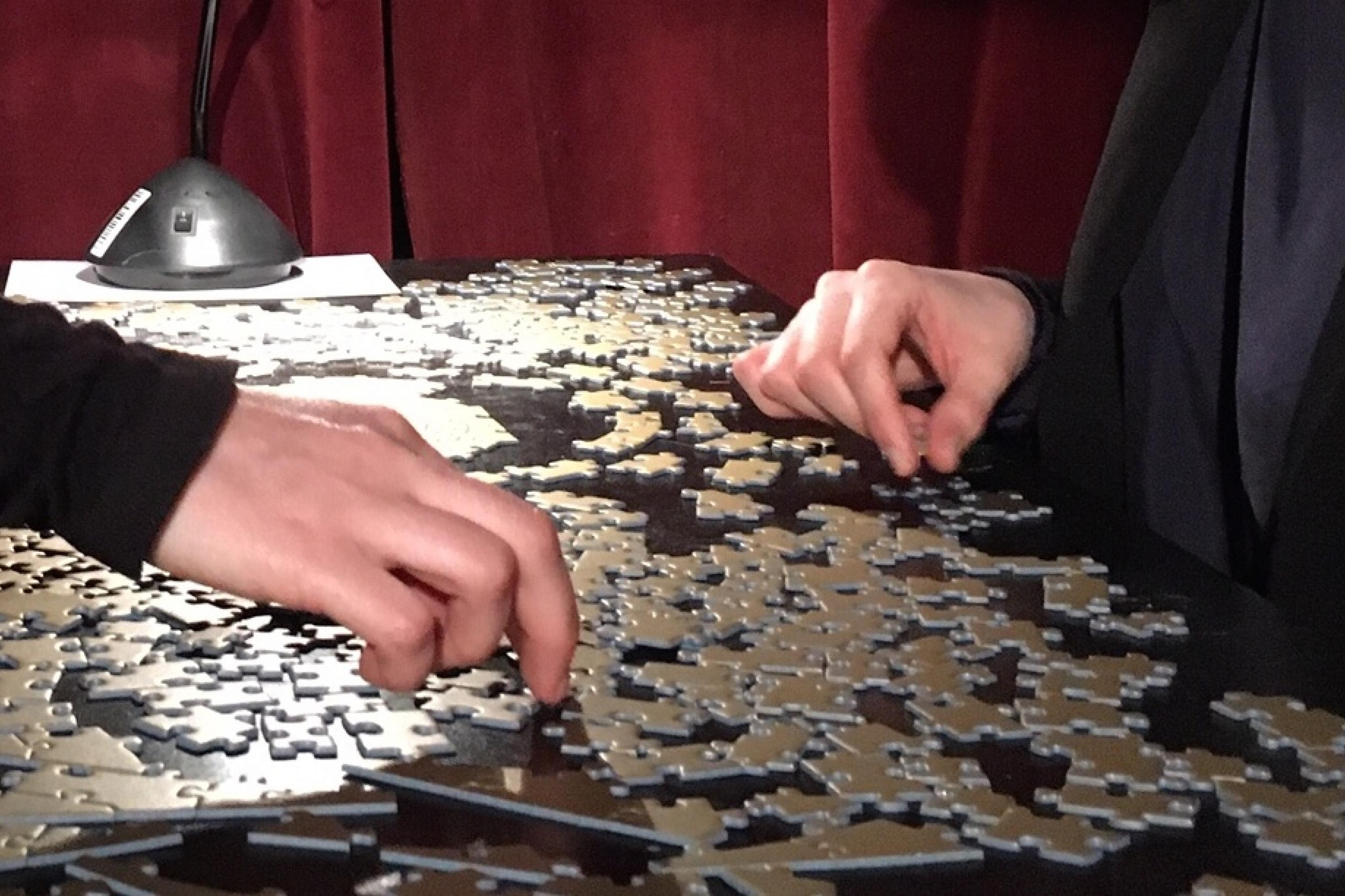 Zwei Menschen sitzen sich gegenüber und puzzlen gemeinsam. Man sieht nur ihre Hände. Die einfarbig goldenen Puzzleteile unterscheiden sich nur durch ihre Form.